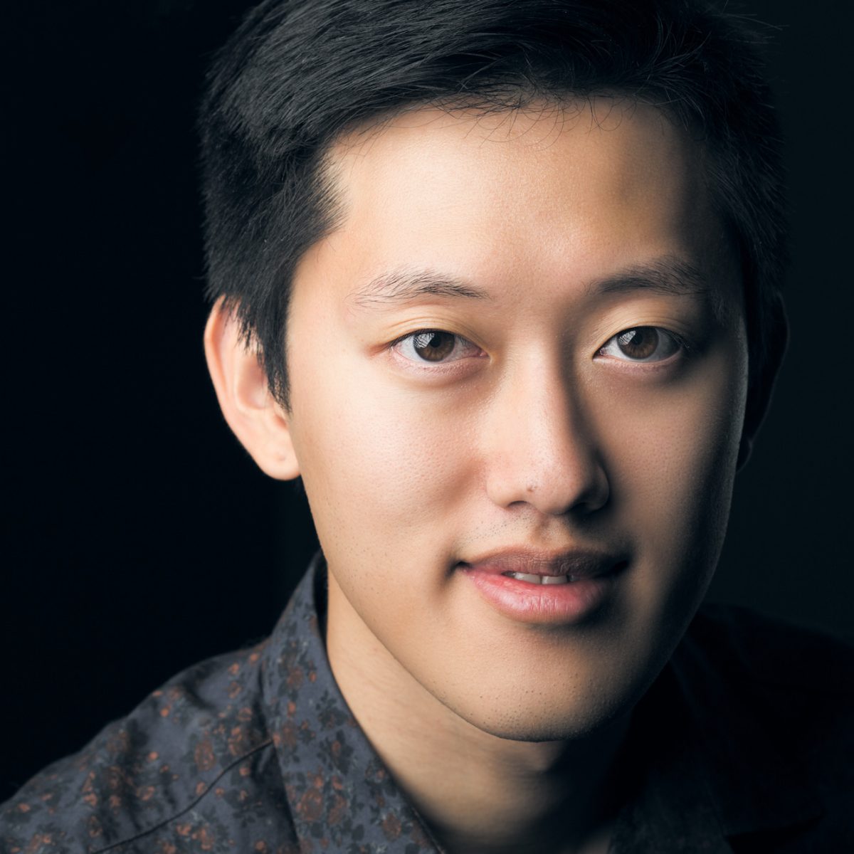 Portrait by Sonoma County Online Dating Profile Photographer: close-up of a young Asian man with short black hair, warm brown eyes, light skin tone, subtle smile, gazing at the camera against a black background, in a dark blue floral-patterned shirt.