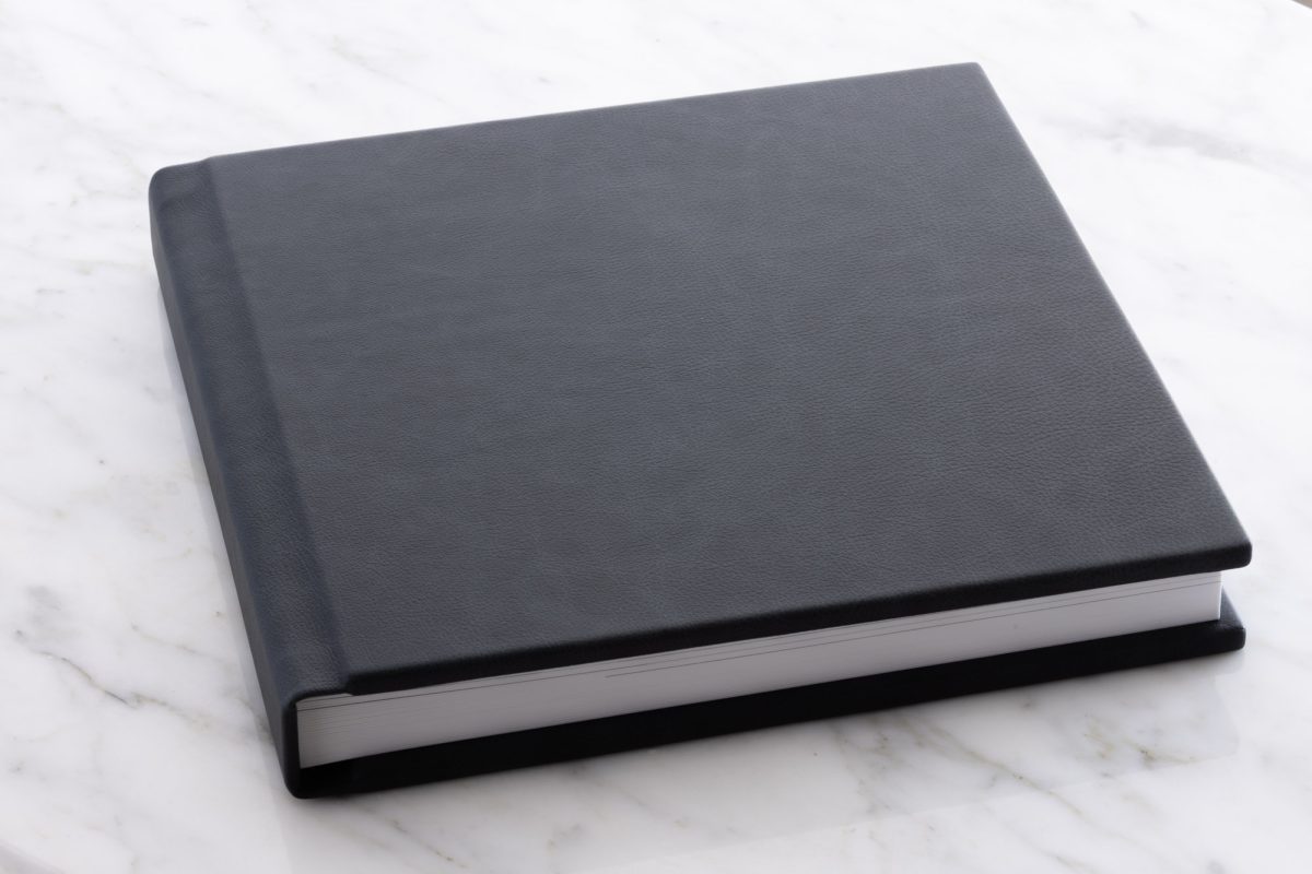 A sleek and elegant engagement album rests closed on a white marble surface, its rich black leather cover exuding a sense of luxury and sophistication. The album has a thick spine and finely bound edges, suggesting high-quality craftsmanship and durability. The white pages within are visible along the edge, tightly bound and hinting at premium photographic prints inside. The contrast between the deep black of the album and the bright, polished marble background highlights the timeless and refined design of this keepsake meant to preserve treasured memories.