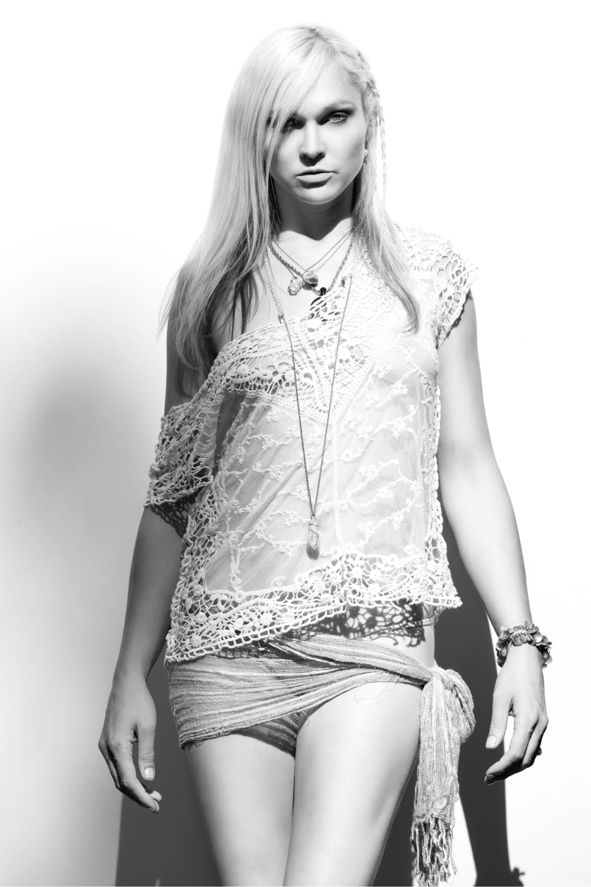 In this high-contrast black and white portrait, a woman stands confidently against a bright white background, her expression calm and direct. She wears a delicate, lace-patterned top with intricate floral detailing and a sheer texture that softly filters the light. Layered necklaces drape down her chest, adding movement and visual interest, while a bracelet adorns her wrist. Her hair is long and straight, with a small braid on one side framing her face. A light wrap is tied at her waist over matching fabric beneath, complementing the airy, bohemian feel of the composition. The clean backdrop and dramatic lighting create a striking silhouette, exemplifying the bold yet refined style of a Calistoga Portrait Photographer.