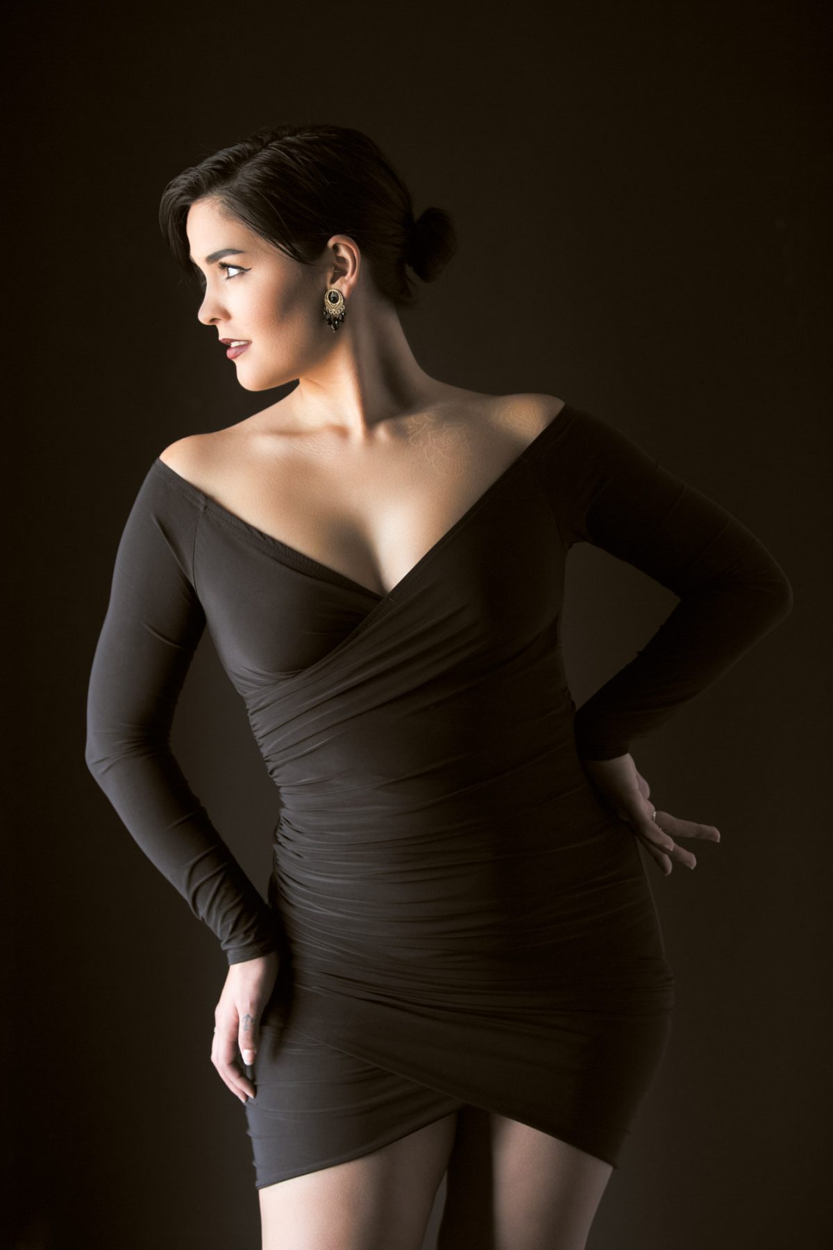 A confident woman poses with elegance against a smooth, dark backdrop, her body slightly angled to the side as she gazes to her left. She wears a sleek, off-the-shoulder black dress that wraps snugly around her torso, accentuating her figure with gentle ruching. The deep neckline highlights her collarbones and the graceful curve of her shoulders. Her dark hair is neatly styled in a low bun, and she wears bold, intricate earrings that add a touch of vintage flair. One hand rests delicately on her upper thigh, while the other is placed behind her back, creating a natural yet sculpted posture. Soft, directional lighting casts a warm glow across her skin, emphasizing the smooth textures and thoughtful composition. The image exemplifies the refined artistry of a Portrait Studio Sonoma County.