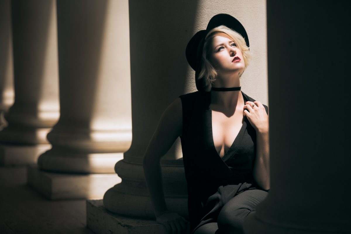 A woman with short blonde hair sits thoughtfully between towering classical columns, her gaze lifted toward a dramatic shaft of sunlight illuminating her face and shoulders. She wears a deep black outfit with a plunging neckline, paired with a black wide-brimmed hat and a velvet choker, creating a striking contrast against the pale stone architecture. Her posture is relaxed yet poised, one hand gently grasping her collar as soft shadows and warm highlights play across her features. The interplay of light and shadow, combined with the timeless elegance of the setting, captures the moody sophistication that defines Bay Area Portrait Sessions.