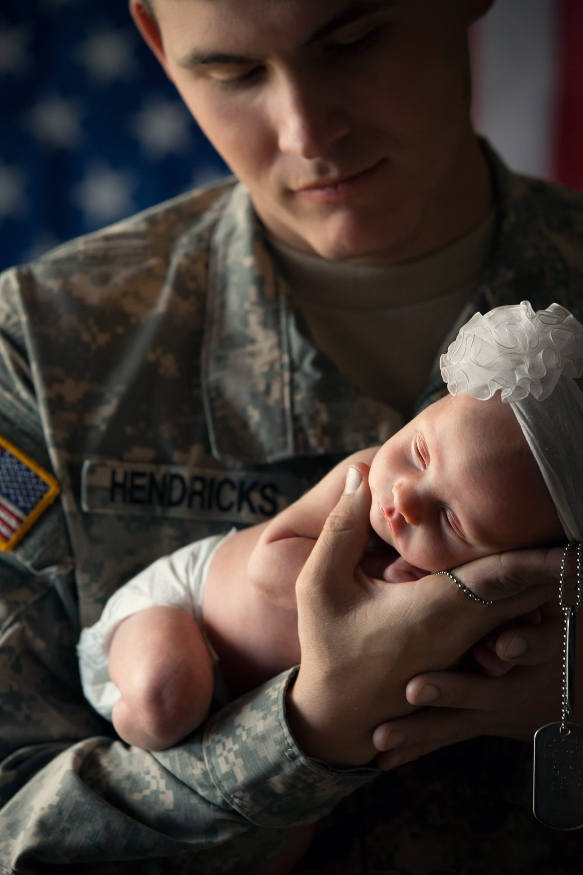 A servicemember dressed in full camouflage uniform cradles a newborn baby gently in both hands, creating a powerful and tender moment. The soldier’s name tag, “HENDRICKS,” and an American flag patch are clearly visible on the uniform, while dog tags hang loosely between his fingers. The infant, swaddled in a white diaper and adorned with a delicate white headband featuring a soft fabric flower, sleeps peacefully in the crook of his arm, her tiny body supported with great care. A softly focused American flag forms the backdrop, adding depth and symbolism to the portrait. The lighting is warm and intimate, casting soft shadows that enhance the emotional weight of the image. This heartfelt and respectful scene exemplifies the poignant storytelling of a Bay Area Military portrait photographer.