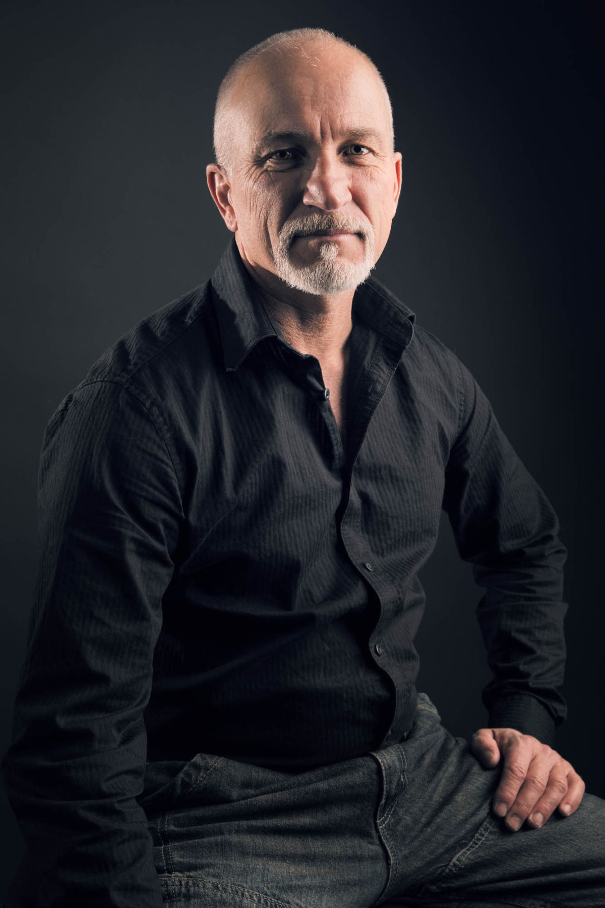 A man with a closely cropped haircut and neatly trimmed white beard sits confidently against a dark, neutral backdrop in this classic studio portrait. He wears a black button-up shirt with a subtle texture and a pair of medium-wash jeans, his posture relaxed yet composed with one hand resting on his thigh. His expression is calm and self-assured, with a hint of a thoughtful smile. The lighting is soft and directional, highlighting the contours of his face and hands while gently fading into shadow on the opposite side, creating depth and character. This refined and grounded composition exemplifies the timeless appeal of Men's Portrait photography Marin County.