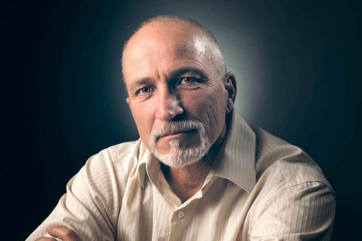 A distinguished man with a neatly trimmed white beard and short-cropped hair is seated against a softly lit, dark gradient background. He wears a cream-colored, pinstriped dress shirt with the collar open, lending a blend of professionalism and relaxed confidence. His arms are gently crossed in front of him, and his gaze meets the camera with a calm, direct presence. The lighting is focused and deliberate, illuminating the contours of his face while subtly fading into the shadows, creating depth and dimension. This thoughtfully composed image exemplifies the clean, refined style of a Professional Portrait photographer Santa Rosa.