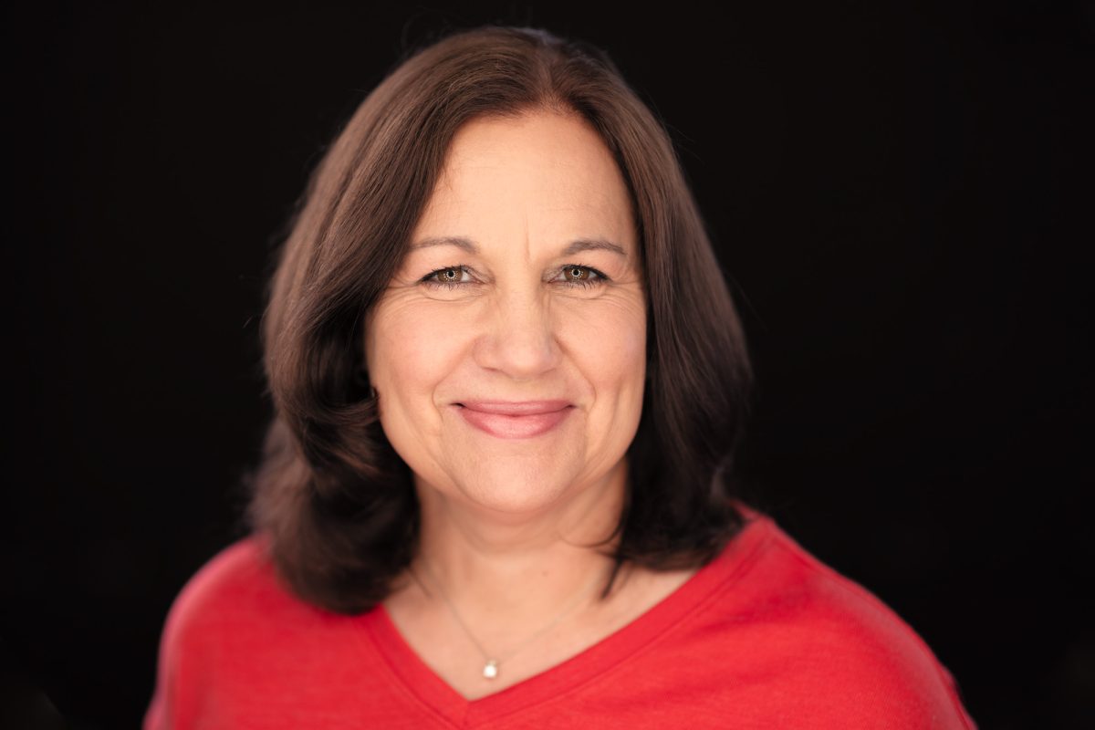 A woman with shoulder-length, dark brown hair and a warm, welcoming smile is photographed against a deep black background. She wears a bright red V-neck top that adds a vibrant pop of color to the composition, along with a delicate silver necklace featuring a small pendant. Her expression is confident and approachable, with soft lighting highlighting her facial features and creating a flattering glow across her skin. Her eyes are gently crinkled at the corners, enhancing the genuine warmth of her presence. This polished and engaging portrait is a strong example of work by a Sonoma County personal branding photographer.