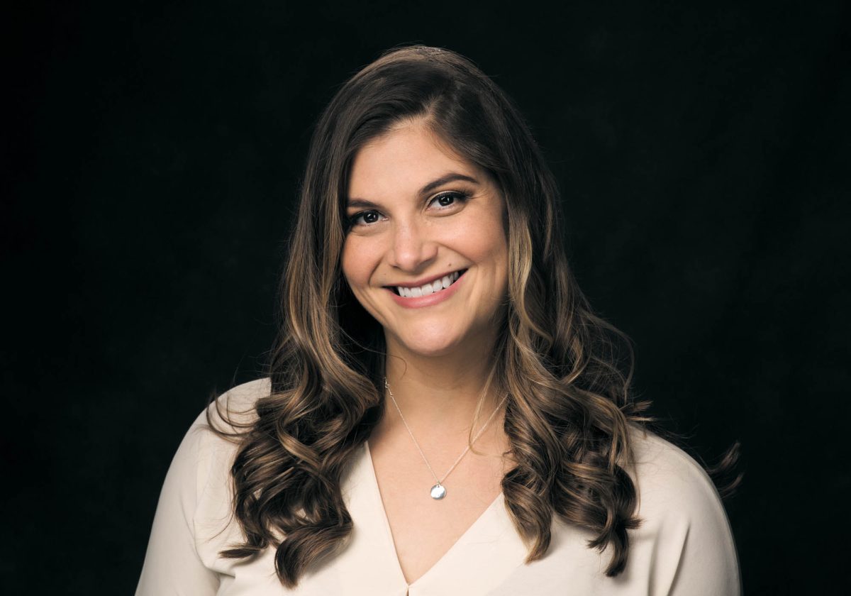 A woman with long, softly curled brunette hair poses in front of a classic dark studio backdrop, wearing a light cream blouse with a modest V-neck and a delicate silver necklace featuring a round pendant. Her expression is open and friendly, with a warm smile and eyes that convey approachability and confidence. The lighting is soft and even, illuminating her face with a gentle glow while allowing natural shadows to add depth and dimension. Her polished appearance and relaxed posture embody the timeless, professional style of Traditional Portrait photography Sonoma county.