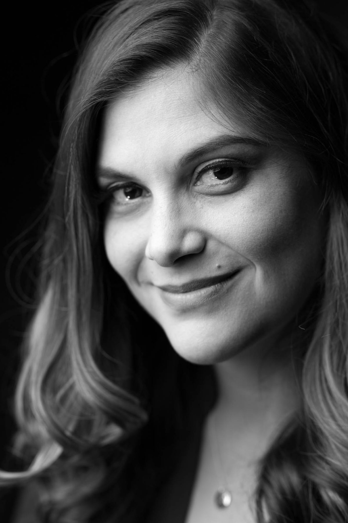 A black-and-white close-up portrait captures a woman with long, softly curled hair as she gazes slightly past the camera with a gentle, knowing smile. The lighting is soft and directional, casting delicate shadows across her face and bringing out the texture of her skin and the natural sheen of her hair. Her eyes are expressive and framed by subtle makeup, and a faint highlight touches the curve of her cheek. A simple round pendant on a fine chain rests at the base of her neck, adding a touch of understated elegance. This timeless, expressive image reflects the refined artistry of a Bay Area portrait photographer.