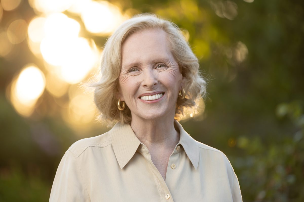A woman with soft blonde hair styled in gentle waves stands outdoors in warm, golden hour light, her expression open and joyful. She wears a light beige button-down shirt and gold hoop earrings, with sunlight softly illuminating her face and hair from behind. The background is a softly blurred blend of greenery and glowing bokeh, creating a serene, natural ambiance. Her relaxed posture and warm smile convey both approachability and wisdom, making this image an ideal example of a Bay Area author portrait photographer capturing authenticity and presence.