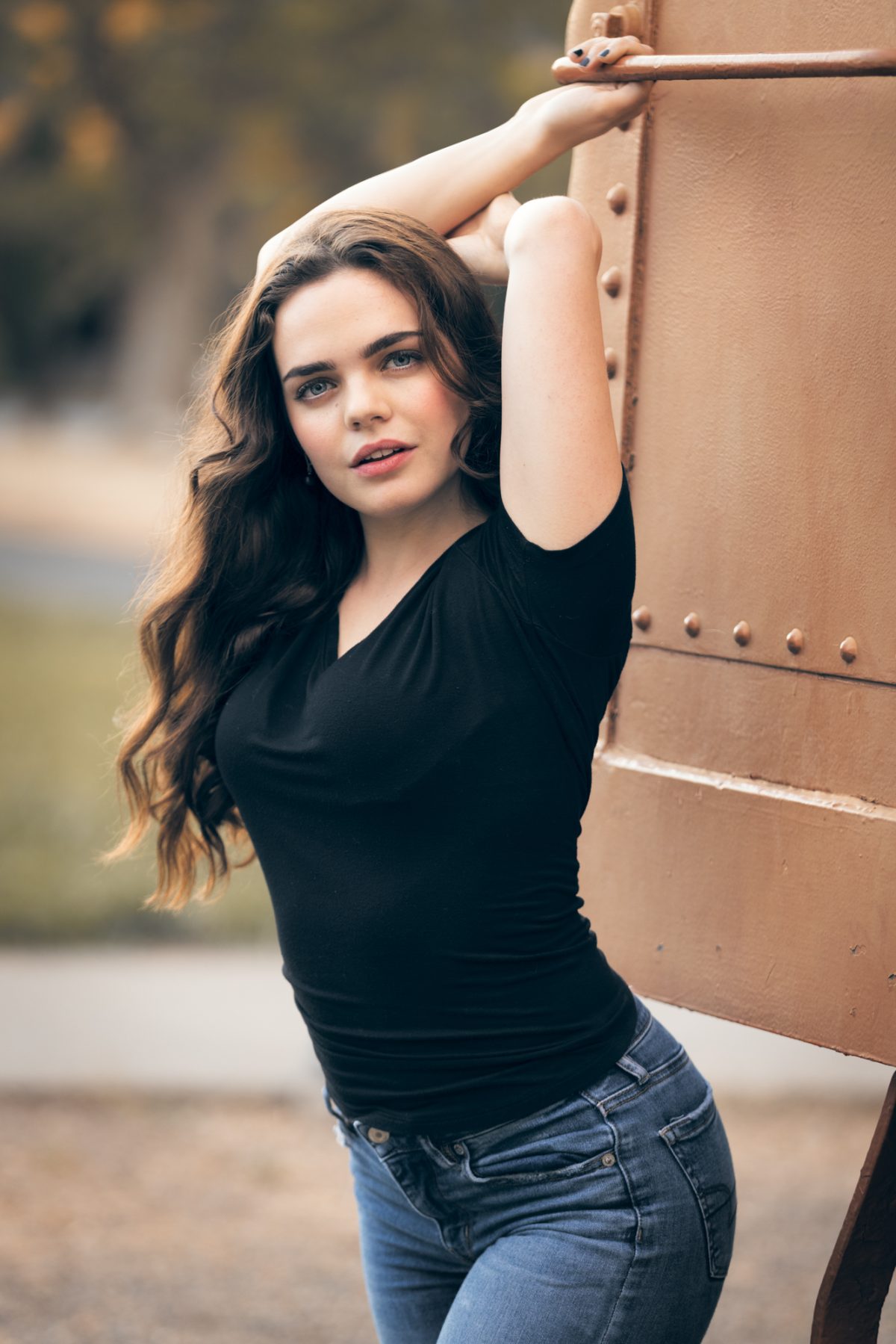 A woman poses confidently outdoors, leaning against a rust-toned metal structure with both arms raised above her head, creating a relaxed and self-assured stance. She wears a fitted black V-neck top and classic blue jeans, highlighting a casual yet polished style. Her long, wavy brown hair cascades over her shoulders, and she gazes directly at the camera with a soft, engaging expression. The background is softly blurred, with hints of greenery and a neutral path, drawing attention to her presence. The natural lighting and approachable styling make this image an ideal example of Professional dating profile photos, showcasing personality, warmth, and confidence.