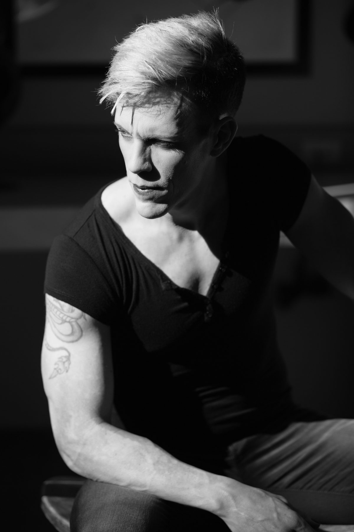 A man sits in dramatic lighting, his figure partially illuminated by a strong shaft of light that defines the contours of his face and muscular build. He wears a fitted black shirt that emphasizes his physique, and his light-colored hair contrasts sharply with the deep shadows surrounding him. A detailed tattoo featuring a coiled snake and a fierce animal head adorns his upper arm, adding a personal element to the otherwise minimalist setting. His expression is serious and contemplative as he looks off to the side, captured in timeless black and white. The careful use of contrast and classic composition embodies the refined aesthetic of Marin County Traditional portraits.