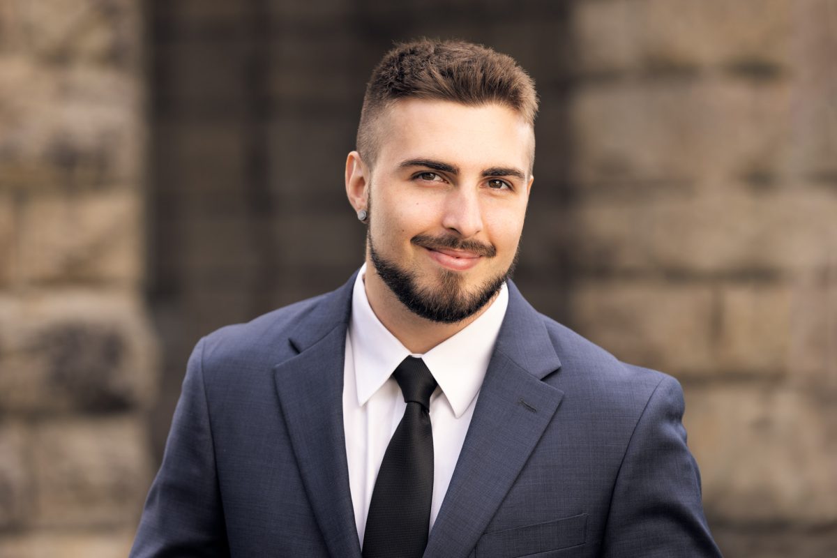 A young man stands confidently in front of a softly blurred stone building, dressed in a sharp dark gray suit with a white shirt and black tie. His neatly groomed beard and short haircut complement his professional appearance, and a subtle smile conveys both approachability and competence. A small earring adds a touch of personal style, while the warm, even lighting enhances his clear complexion and bright eyes. The setting and attire create a polished, trustworthy image ideal for industry professionals. This portrait exemplifies the refined, business-ready style of a real estate portrait photographer.
