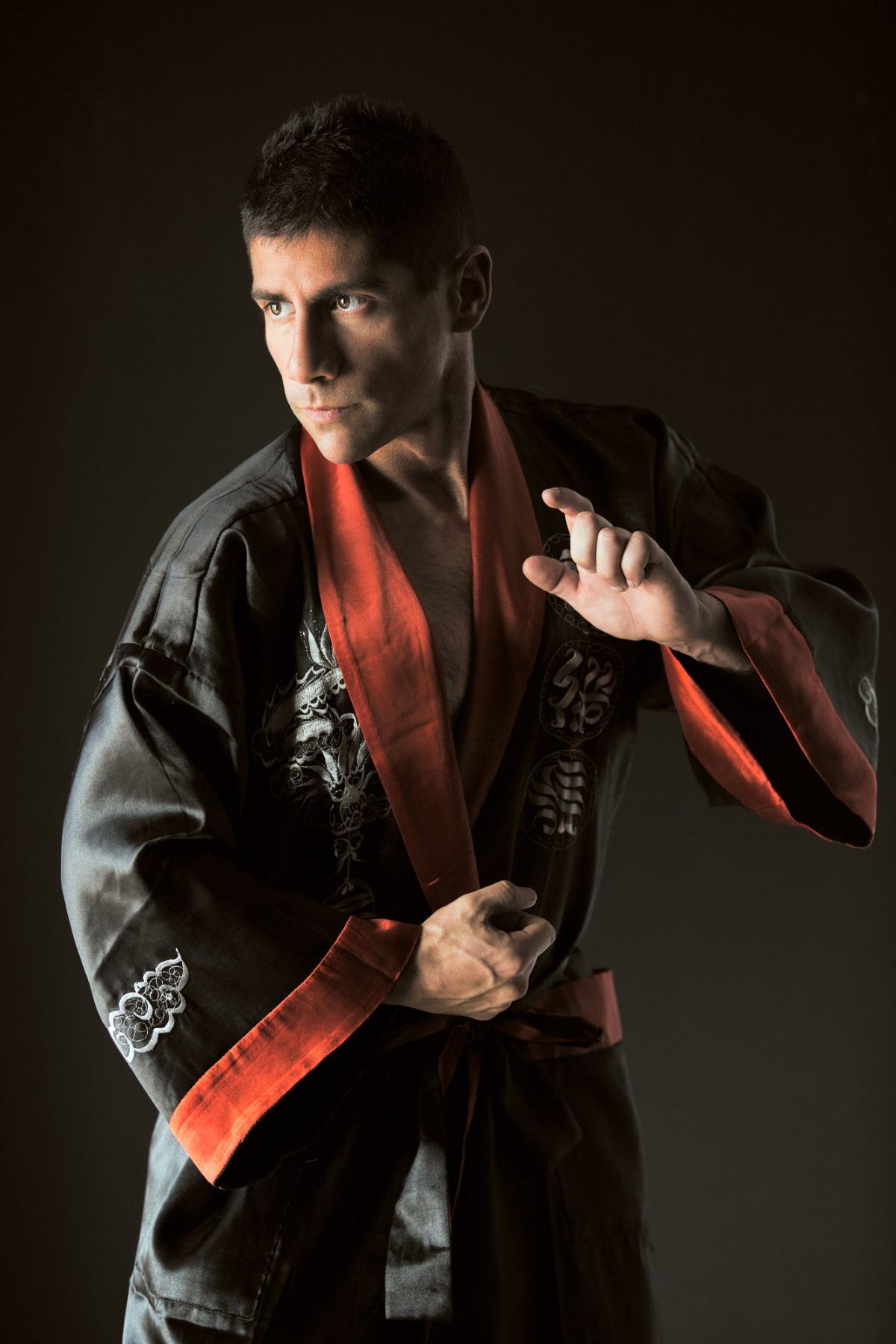 A man stands in a poised martial arts stance, wearing a black satin robe with bold red trim and intricate silver embroidery along the sleeves and chest. His left hand is drawn tightly at his waist, gripping the robe’s belt, while his right hand is raised with fingers delicately curved in a controlled, focused gesture. His gaze is intense and directed slightly to the side, emphasizing concentration and readiness. Dramatic lighting highlights the contours of his face and the sheen of the fabric, casting soft shadows that enhance the depth and texture of the scene. The neutral, shadowed background keeps the focus on his expression and form, creating a powerful and disciplined image that reflects the refined strength of martial arts portraits Northern California.