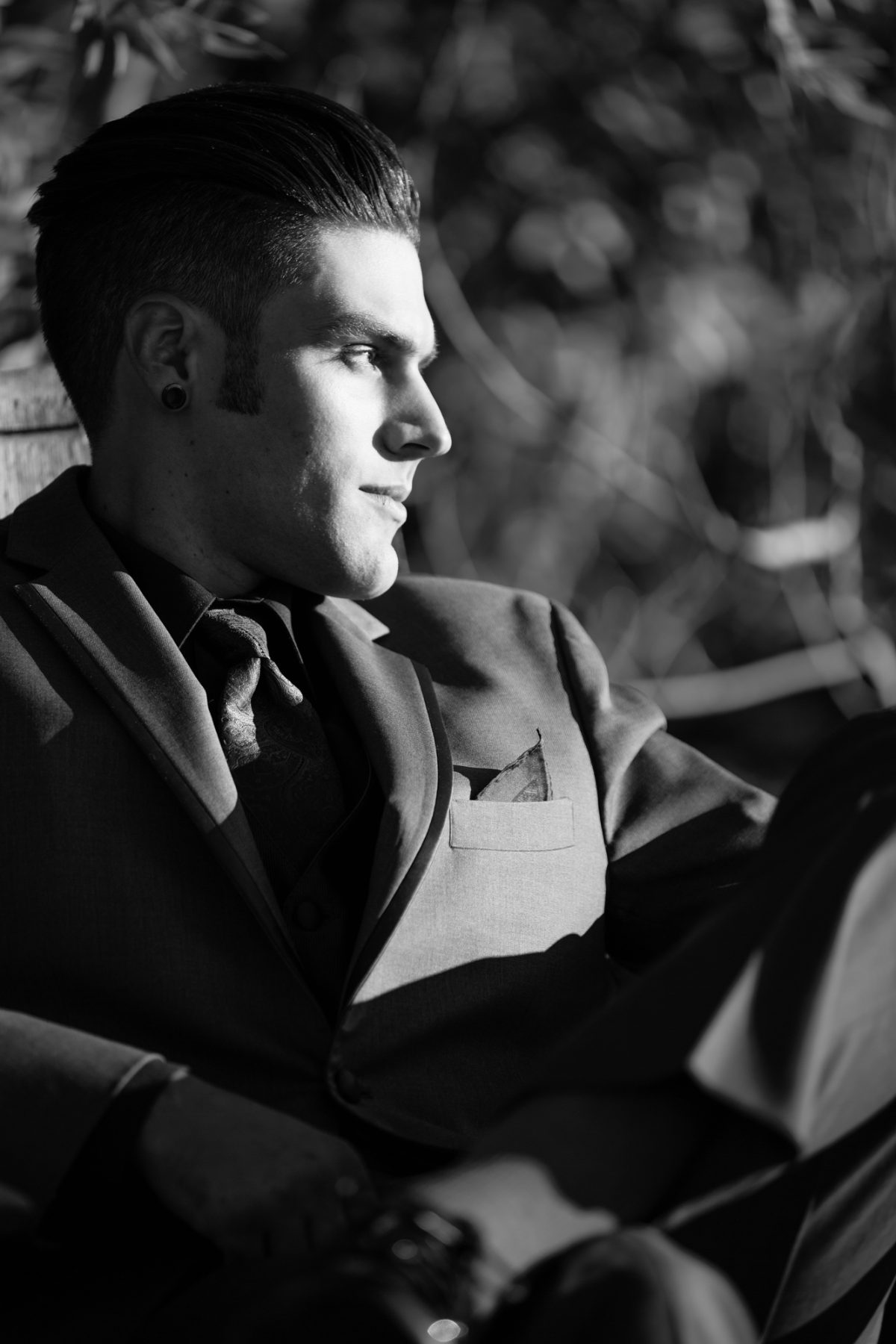 A man sits in thoughtful repose, dressed in a tailored suit with a pocket square and patterned tie, his posture relaxed yet composed. Captured in dramatic black and white, the image uses strong directional light to highlight the sharp lines of his jaw, the smooth structure of his suit, and the confident styling of his slicked-back hair. His gaze is directed off-frame, adding a contemplative depth to the portrait. Subtle background blur and shadowed foliage provide texture without distraction. This powerful and refined image exemplifies the character-driven elegance of Men's Portraits Sonoma County.