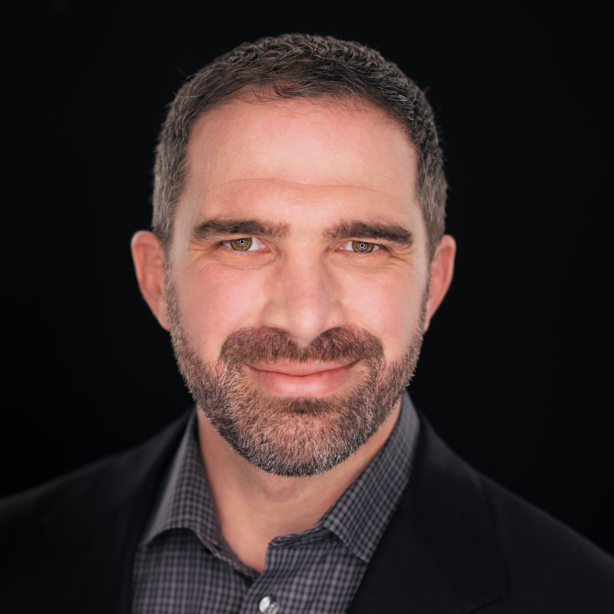 A professional headshot of a man with short, neatly trimmed salt-and-pepper hair and a full beard, smiling warmly at the camera. He has light skin and expressive hazel eyes that reflect the studio lighting. He is dressed in a dark blazer over a checkered button-down shirt, with the top button casually undone. The background is a solid black, providing contrast that draws focus to his face. The lighting is soft and even, highlighting the texture of his beard and the detail in his shirt. This image features Family Photographer Jason Guy prominently, presenting a friendly and approachable demeanor ideal for his professional role.