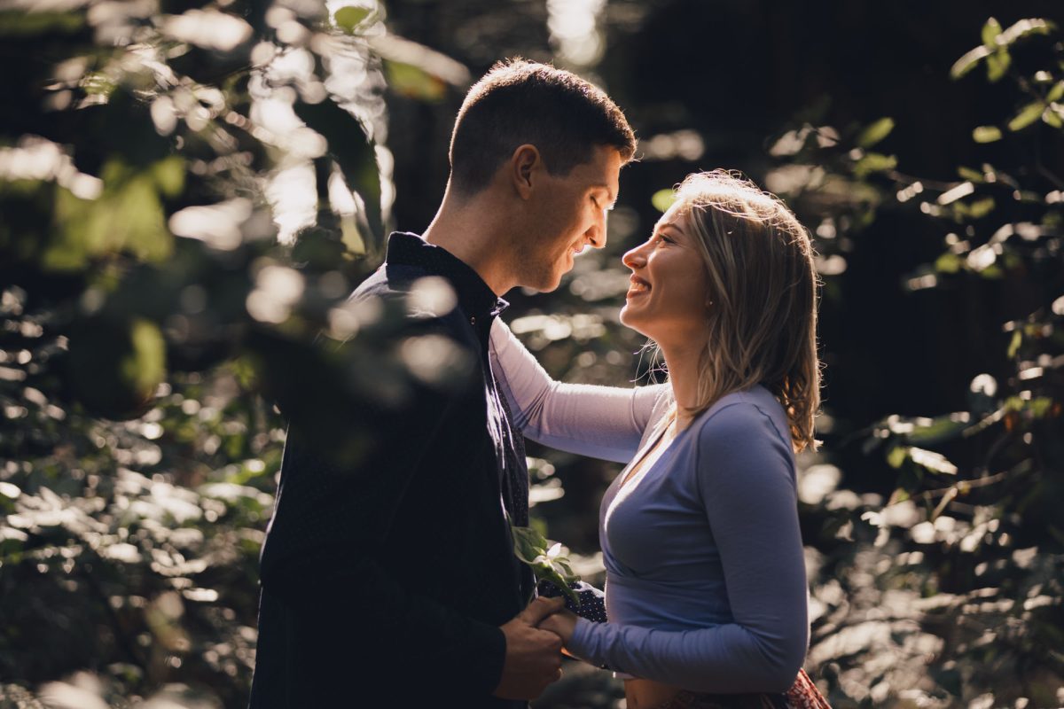 A couple stands in a sun-dappled forest clearing, their faces close and glowing with happiness as they gaze into each other's eyes. The man, dressed in a dark button-up shirt, smiles tenderly as he holds the woman's hands. She wears a soft lavender long-sleeved top and a patterned skirt, her expression joyful and affectionate. A small green stem peeks from their clasped hands, adding a natural touch to the moment. Gentle sunlight filters through the surrounding leaves, creating a romantic, serene atmosphere that beautifully captures the essence of an Engagement Photographer Sonoma County session.