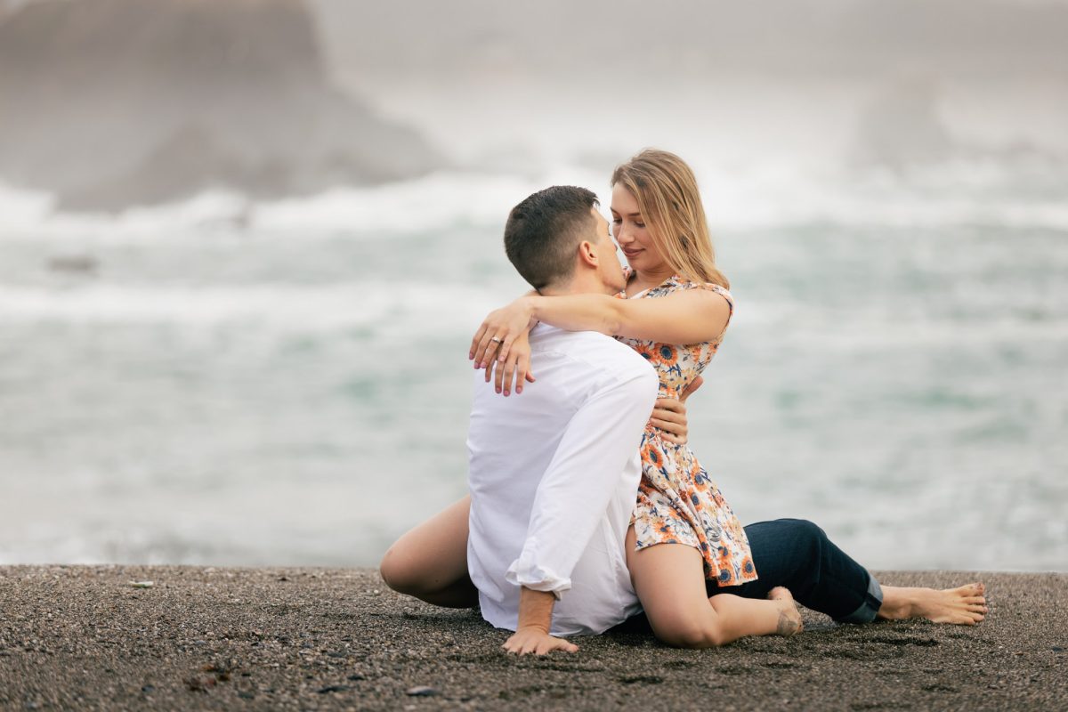 A couple sits closely together on the shoreline, embraced in a tender moment as waves roll gently in the background. The man, wearing a crisp white shirt and dark rolled-up pants, leans back slightly with one hand supporting him in the sand, while the woman kneels beside him in a short, floral dress, wrapping her arms around his shoulders. Their faces are near, eyes softly closed or gazing at one another, lost in their connection. The misty sea air blurs the rocky coast behind them, creating a romantic, intimate mood that beautifully reflects the essence of Sonoma County Couples Portrait Sessions.
