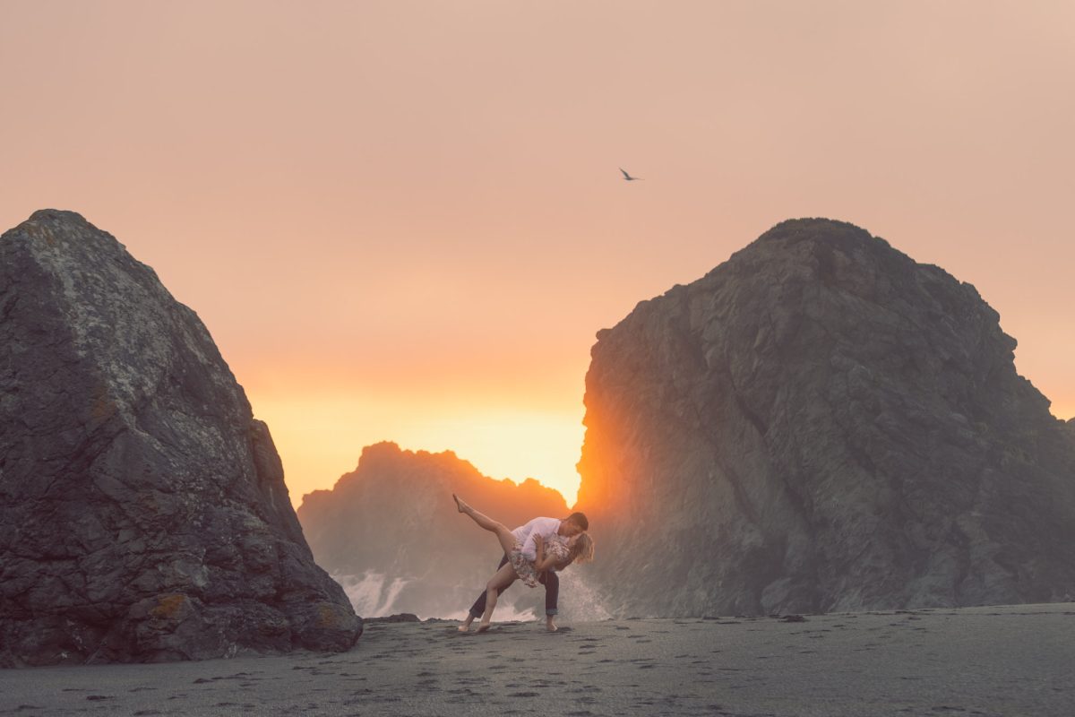 A romantic moment unfolds on the shore as a couple dances barefoot in the sand, framed by massive sea stacks and the fiery glow of a setting sun. The man gently dips the woman backward in a graceful pose, her leg extended in midair, their silhouettes outlined against the warm golden light. Waves crash softly behind them, while a single bird glides across the sky above. The rugged textures of the dark coastal rocks contrast with the tender energy between the couple, capturing a timeless sense of connection and natural beauty that defines Couples portraits Bodega Bay.