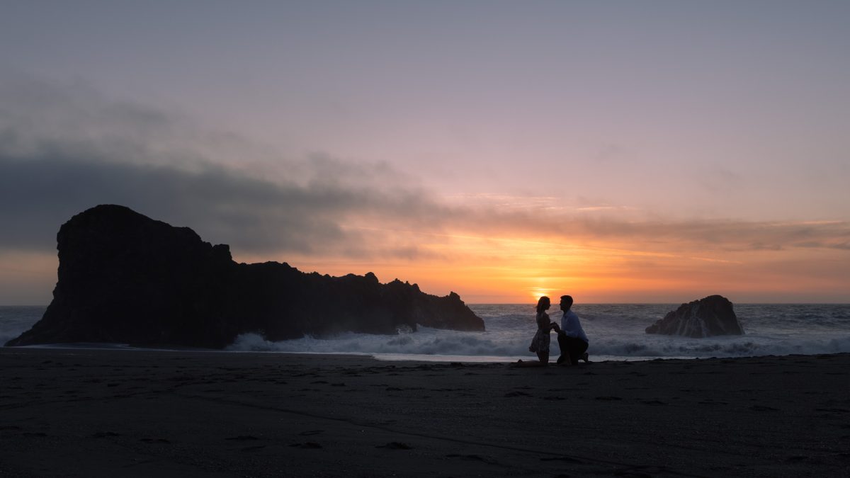 A couple is silhouetted against a breathtaking coastal sunset, kneeling face-to-face on a dark sandy beach as waves crash gently behind them. The man appears to be proposing, holding the woman's hands tenderly while they share a quiet, intimate moment. The sky is painted in soft hues of lavender, peach, and gold, casting a warm glow along the horizon. Jagged sea rocks rise dramatically from the water, framing the couple within the natural landscape. The scene is peaceful, romantic, and timeless—perfectly capturing the emotion and setting of engagement portraits Sonoma County.