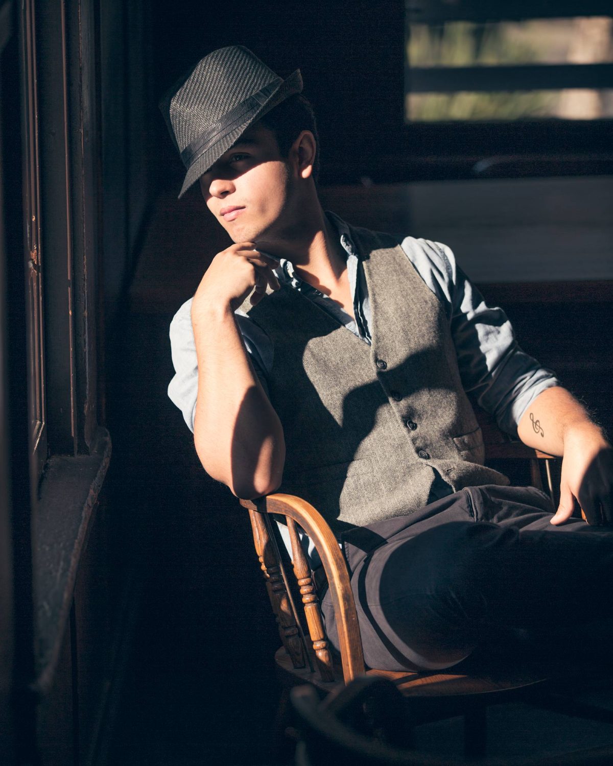 A man sits in a wooden chair by a window, partially bathed in warm, directional sunlight that casts dramatic shadows across his face and body. He wears a textured gray vest over a rolled-sleeve button-down shirt, paired with dark pants, and tilts a pinstriped fedora down to partially veil his eyes, adding a sense of mystery and sophistication. His relaxed posture and the thoughtful hand resting under his chin convey quiet confidence. A small tattoo is visible on his inner forearm, hinting at personal style beneath the classic look. This stylish and intimate composition reflects the refined character of Sonoma County Men's Portraits.