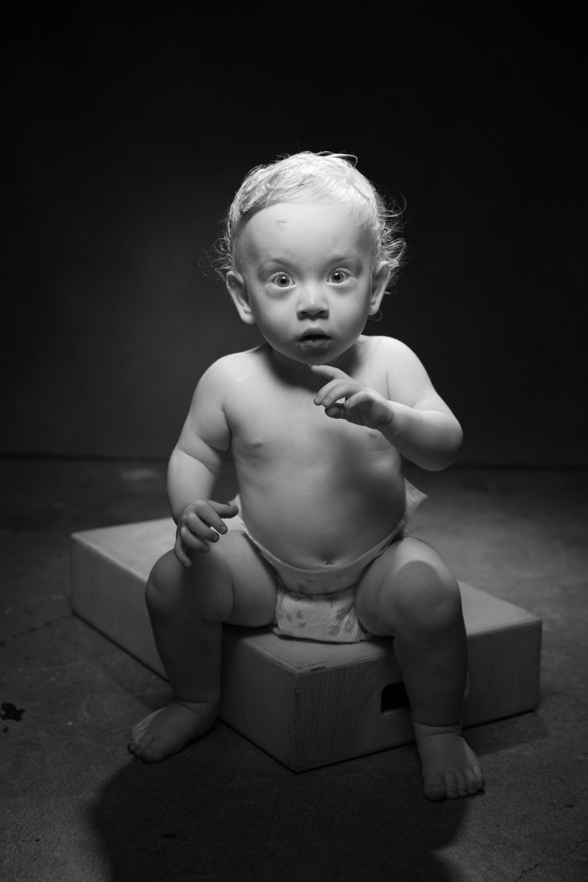 A young child sits on a wooden box in the center of a dimly lit studio, illuminated by a single overhead light that casts dramatic shadows around the scene. The child, wearing only a patterned diaper, is captured mid-gesture with one hand raised and wide eyes looking directly toward the camera in an expression of surprise or curiosity. Their curly, light hair catches the light, creating a halo effect that contrasts with the dark backdrop and floor. The textured studio setting and the use of black and white photography give the image a classic, timeless quality. This expressive and artfully composed moment reflects the creativity and character of Children's portrait sessions Sonoma County.