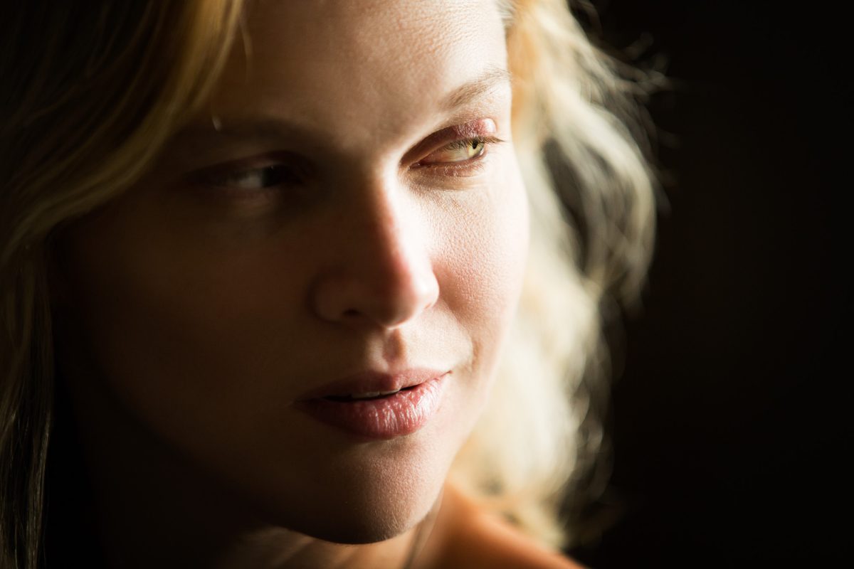 A close-up portrait captures a woman in soft, natural light as she gazes off to the side with a thoughtful expression. The composition highlights the texture of her skin and the delicate details of her face—her gently parted lips, light lashes, and softly lit cheekbones. Her blonde hair falls loosely around her face, with strands catching the ambient light and adding depth to the background, which fades into a dark blur. The shallow depth of field and subtle glow create a quiet, introspective mood, reflecting the artistic sensitivity of a Petaluma portrait photographer.