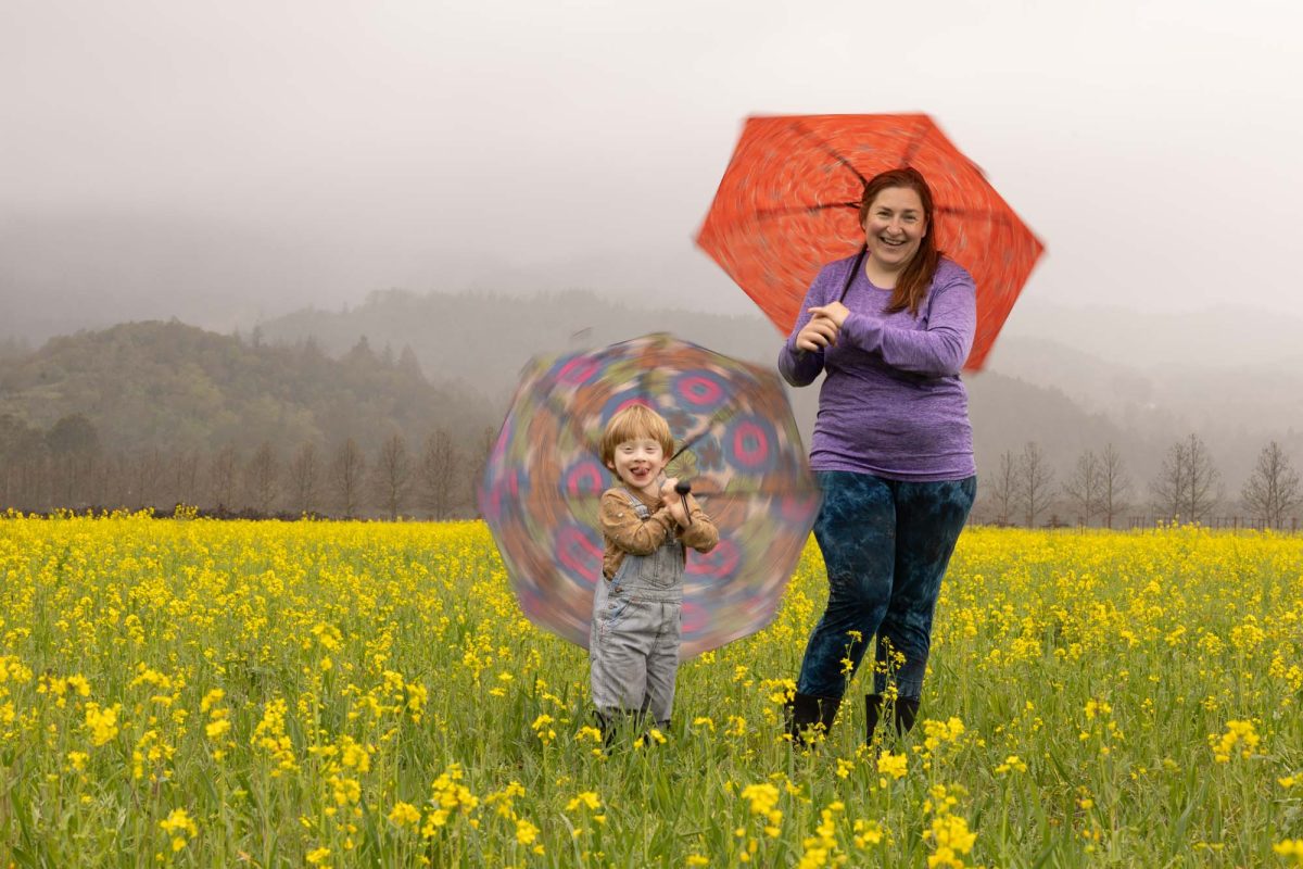 A woman and a young child stand in the midst of a vibrant field of yellow wildflowers, both holding umbrellas on a misty, overcast day. The child, dressed in striped overalls and a tan shirt with playful prints, beams with excitement while spinning a clear umbrella adorned with colorful circular patterns, creating a slight blur of motion. Beside the child, the woman, wearing a purple long-sleeved top and dark patterned leggings, smiles warmly beneath a red-orange umbrella. Behind them, rows of bare trees and mist-covered hills add depth and mood to the scene. The joyful interaction and lush floral setting highlight the natural charm and playfulness that define Marin County Wild Flower Family Portrait sessions.
