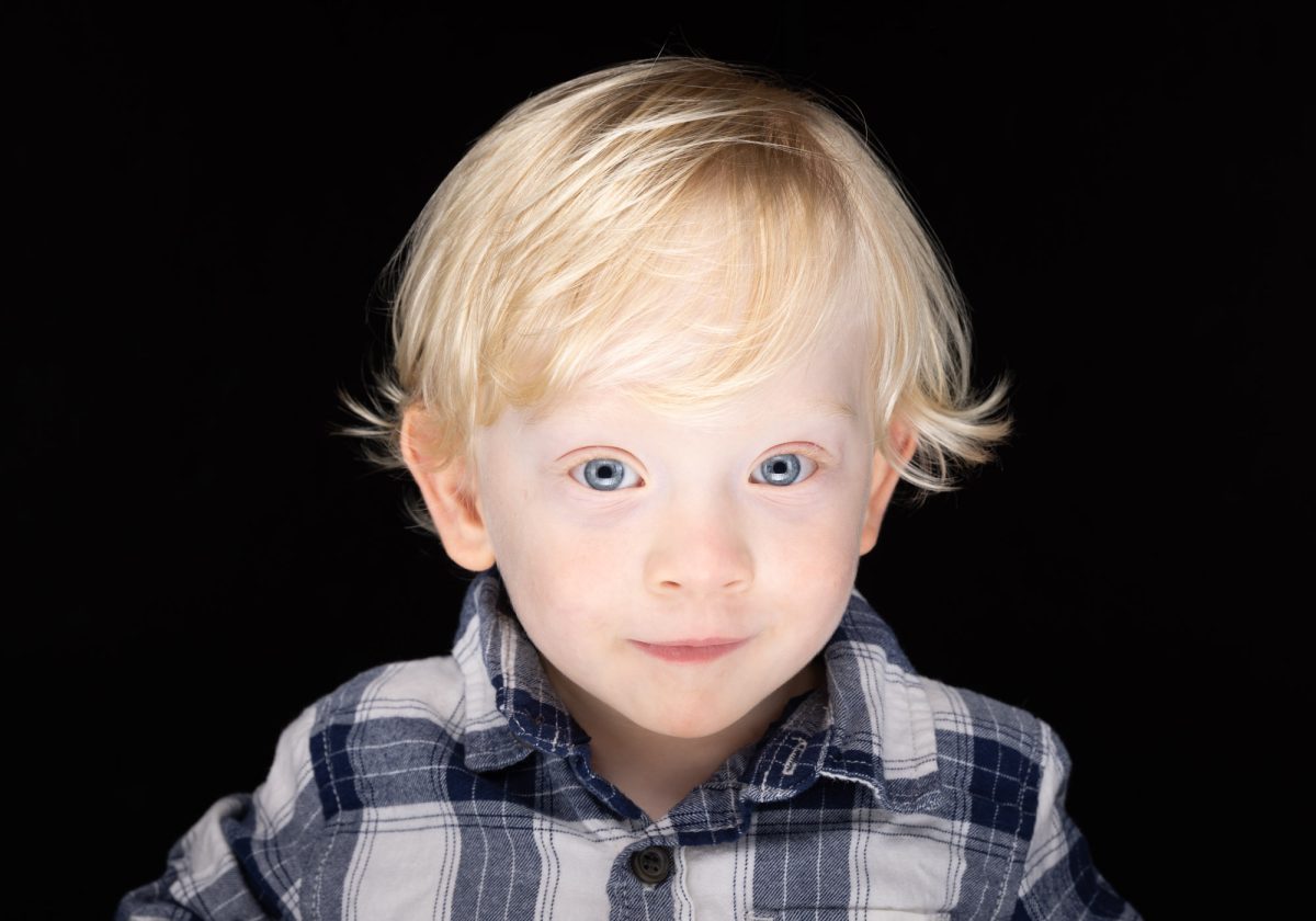 A young child with bright blue eyes and tousled platinum blonde hair stands against a deep black background, gazing directly at the camera with a soft, inquisitive expression. The child wears a blue and white plaid button-up shirt, the collar slightly open and the fabric softly creased, adding a casual and natural feel. Even lighting highlights the delicate features of the child's face, from the subtle pink of the cheeks to the light lashes and brows, giving the portrait a clean, polished look. The dark backdrop contrasts beautifully with the child’s light hair and fair complexion, drawing full focus to the eyes and gentle smile. This timeless, expressive image captures the heartwarming simplicity of Sonoma County children’s portrait sessions.