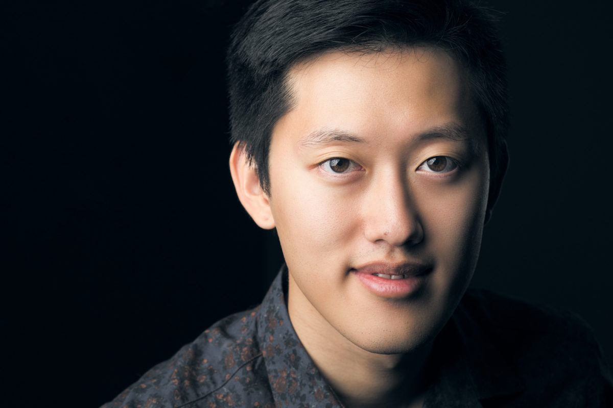 A young man is photographed in a close-up portrait against a dark, solid background. He is slightly angled toward the camera with a gentle, confident expression and soft smile. His short, neatly styled black hair frames his face, and the lighting highlights the smooth texture of his skin and the natural contours of his features. He wears a dark button-up shirt with a subtle floral pattern, which contrasts softly with the background. The overall composition, focus, and lighting emphasize clarity and professionalism, exemplifying the skill of a Headshot photographer Sonoma County.