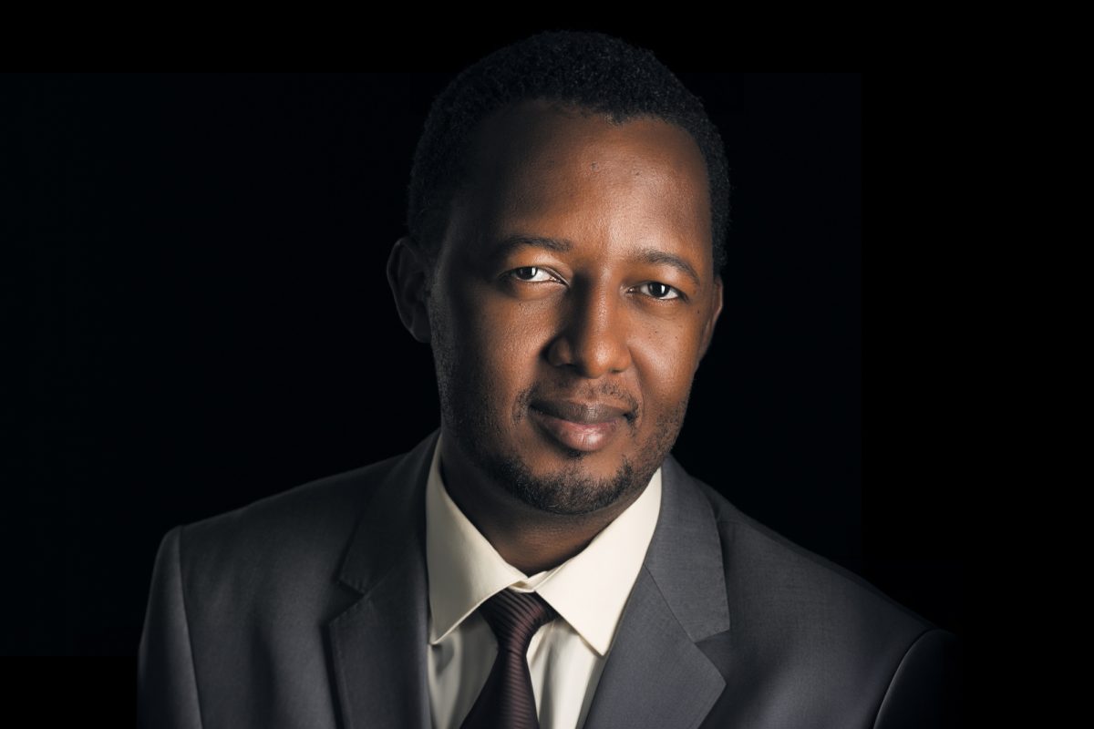 A professionally composed headshot of a man set against a solid black background, creating a strong contrast that emphasizes the subject. The man has short, neatly trimmed black hair and a closely groomed beard. He wears a medium-gray suit with a white dress shirt and a dark brown tie. His expression is confident and approachable, with a gentle smile and direct eye contact with the camera. The lighting is soft and directional, highlighting the right side of his face while gradually fading into shadow on the left, enhancing the contours and depth of his features. This image represents the refined, polished style associated with Sonoma County Professional Headshots.