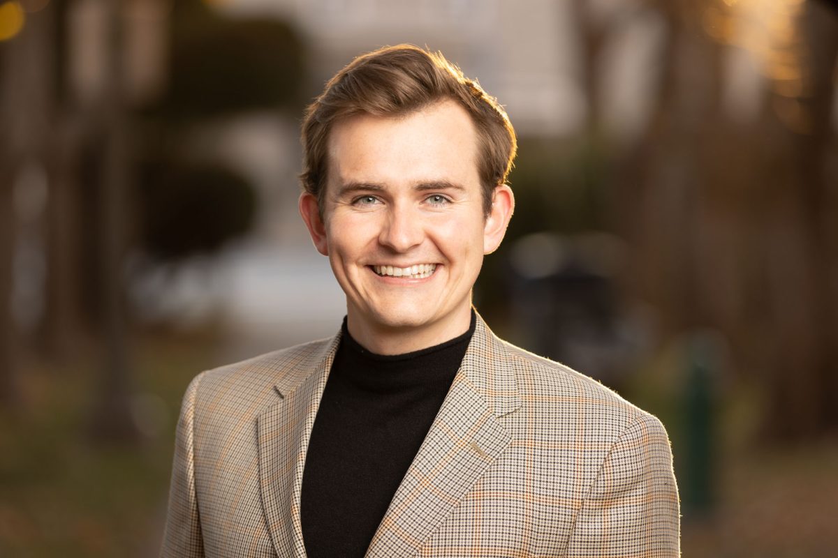 A professionally composed outdoor headshot of a young man standing in soft, golden hour lighting. He smiles warmly, looking directly at the camera with bright eyes and a relaxed, confident expression. His light brown hair is neatly styled, and his skin tone glows under the gentle sunlight. He wears a light brown plaid blazer over a black turtleneck, combining classic and contemporary style elements. The background features a softly blurred residential street lined with tall trees and muted seasonal foliage, creating a warm, natural ambiance that enhances the subject’s professional appearance. This image exemplifies the polished, approachable style found in Professional headshots Sonoma County.
