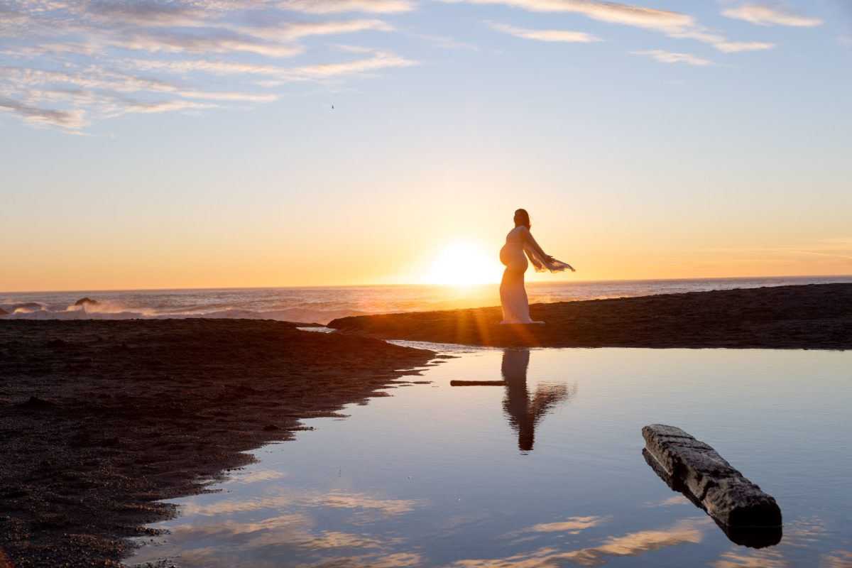 A pregnant woman in a flowing white dress stands gracefully with arms gently extended on a dark rocky outcrop overlooking the Pacific Ocean during a Sonoma County maternity photo shoot. Golden hour sunlight from the setting sun creates a brilliant backlit silhouette and radiant lens flare while her clear reflection shimmers in a shallow tide pool in the foreground as waves with white foam roll toward the shore under a sky blending soft blue and warm orange tones.