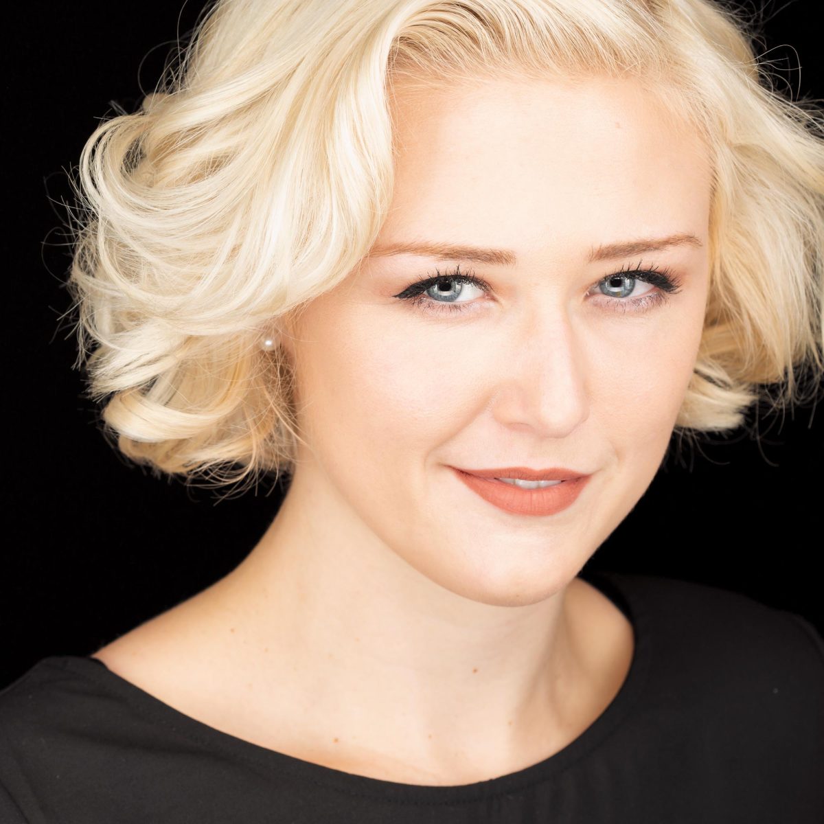 A young woman with platinum blonde, softly curled hair poses in a high-resolution headshot against a seamless black background. Her hair is styled in voluminous waves that frame her face, with a side part adding dimension. She has light blue eyes accentuated by well-defined eyeliner and mascara, and her warm-toned lipstick complements her fair complexion. A subtle pearl earring is just visible beneath her hair. She wears a simple, elegant black top with a rounded neckline. The lighting is bright and evenly distributed, creating a polished and professional image that highlights the clarity and precision typical of Sonoma County Modeling Headshots.