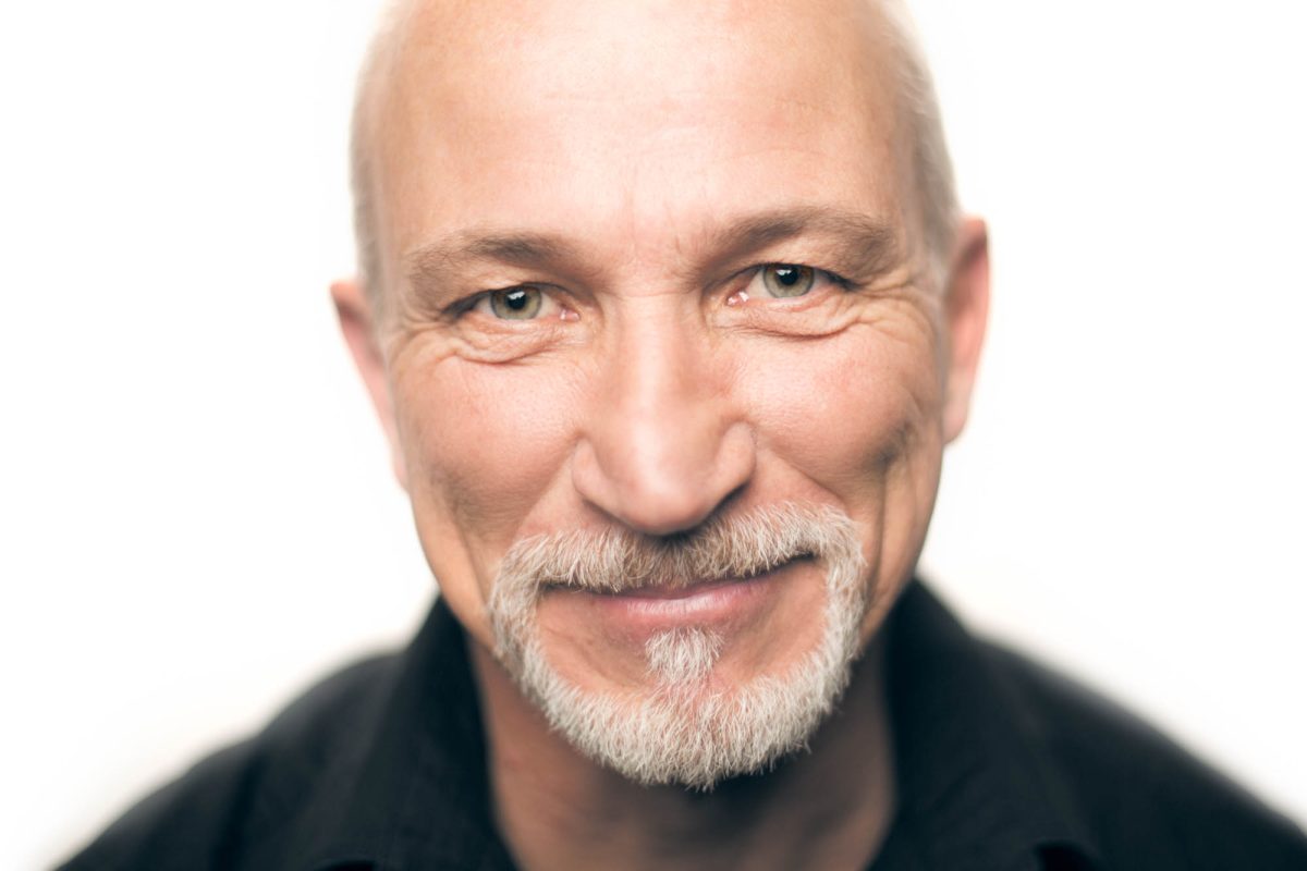 A mature man with a bald head and neatly groomed white goatee is photographed in a warmly lit close-up portrait. His expression is kind and approachable, with a gentle smile that reaches his light green eyes, which are framed by natural lines and slight crow's feet. He wears a dark collared shirt that contrasts subtly against the soft, high-key white background. The image is sharply focused on his face, with a shallow depth of field that brings attention to his expression and features. This image is a refined example of Professional headshots Sonoma County.