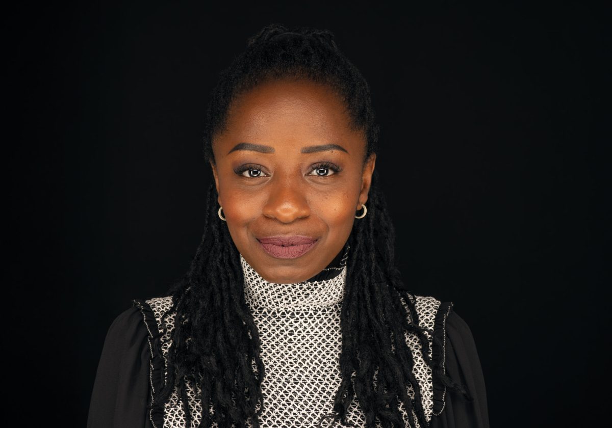 A confident woman stands centered against a solid black background, facing the camera directly with a poised expression. Her dark, tightly coiled locs are pulled back from her face, cascading over her shoulders. She wears a high-necked blouse featuring a geometric black-and-white pattern on the front panel, complemented by black sleeves. Her makeup is natural yet defined, with full brows, long lashes, and mauve lipstick. Gold hoop earrings add a subtle accent. The lighting is soft and even, highlighting her skin tone and facial features. This professional portrait exemplifies the work of a Sonoma Headshot Photographer.