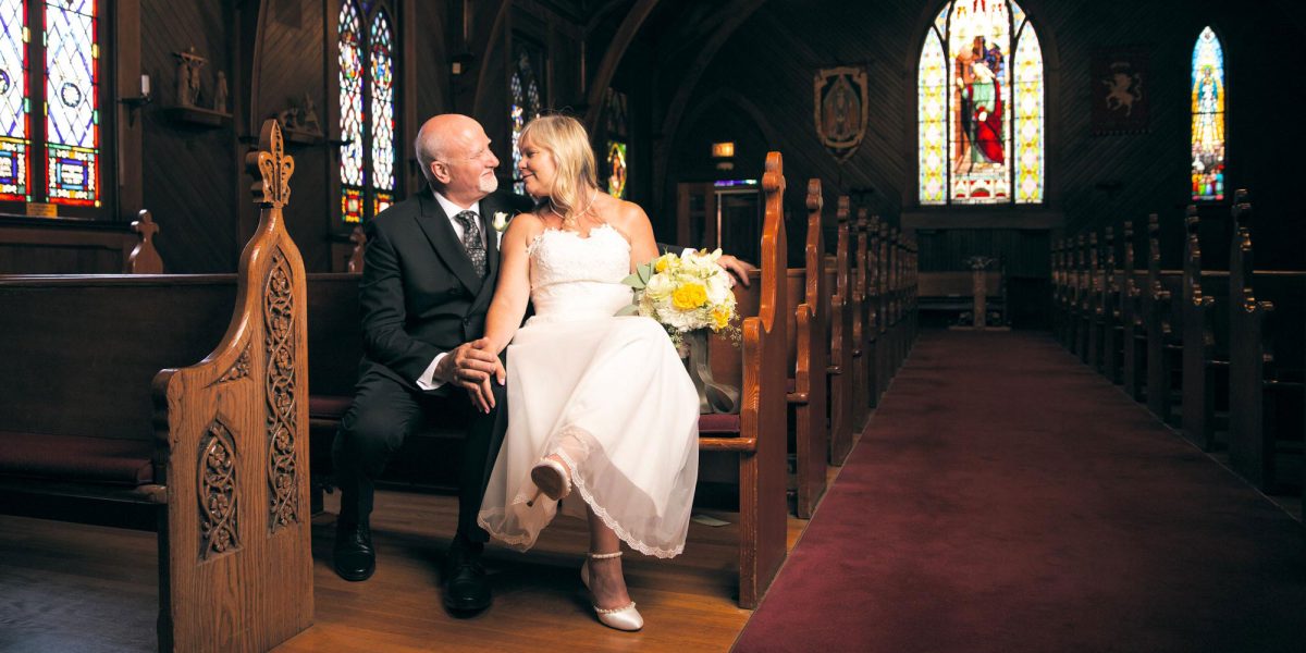 In a warmly lit, traditional wooden church adorned with arched ceilings and glowing lanterns, a bride and groom sit affectionately side by side on a pew near the front aisle. The bride, dressed in a sleeveless white gown with lace detailing and a tea-length hem, holds a vibrant bouquet of yellow and white flowers with greenery. She wears white ankle-strap heels and gazes lovingly at her partner. The groom, in a dark suit with a patterned tie and boutonnière, gently clasps her hand and smiles back at her. Ornate stained glass windows illuminate the sanctuary with vivid colors and religious imagery. The polished wooden pews and intricate carvings enhance the sacred ambiance. This tender moment captures the joy and intimacy of a Chruch of the Incarnation Wedding.