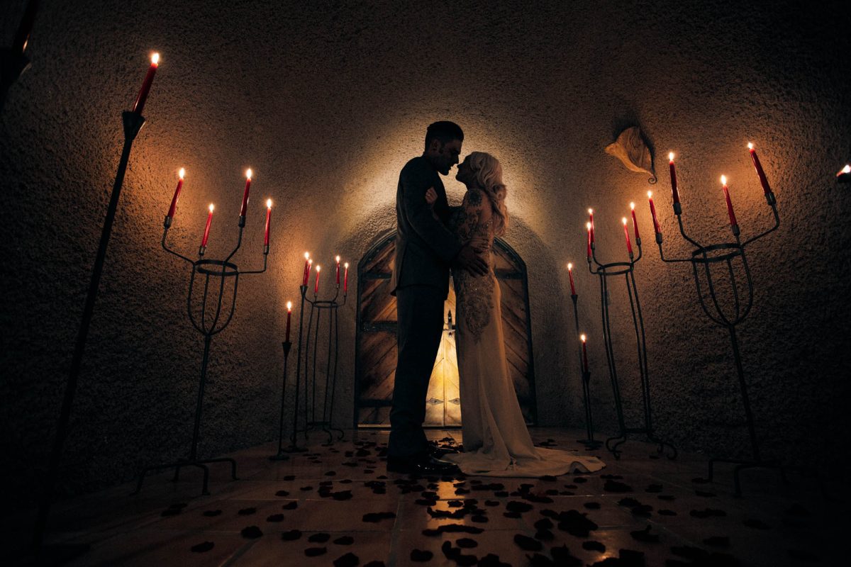Sonoma wine cave wedding featuring a romantic, low-lit scene captures a bride and groom standing face to face in an intimate stone hallway illuminated by tall candelabras holding red taper candles. The couple is framed by an arched wooden door behind them, which glows softly with backlighting that creates a warm halo effect around their silhouettes. The bride wears a fitted, floor-length gown with sheer sleeves and intricate embellishments, while the groom is dressed in a classic suit. Scattered rose petals line the tiled floor, adding texture and deep red accents to the dimly lit space. This atmospheric and moody moment beautifully exemplifies Sonoma Valley Wedding Photography.