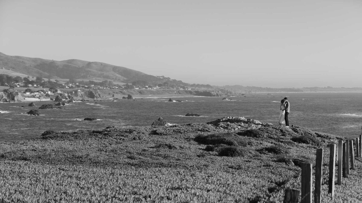 A black and white landscape photograph from Duncan's Landing couple's photos, capturing a romantic moment where a couple embraces on a rugged, grassy cliffside overlooking the vast Pacific Ocean. Gentle waves crash against rocky outcrops below, while a distant coastal town nestles amid rolling hills under a clear sky, evoking a sense of serene isolation and timeless love.