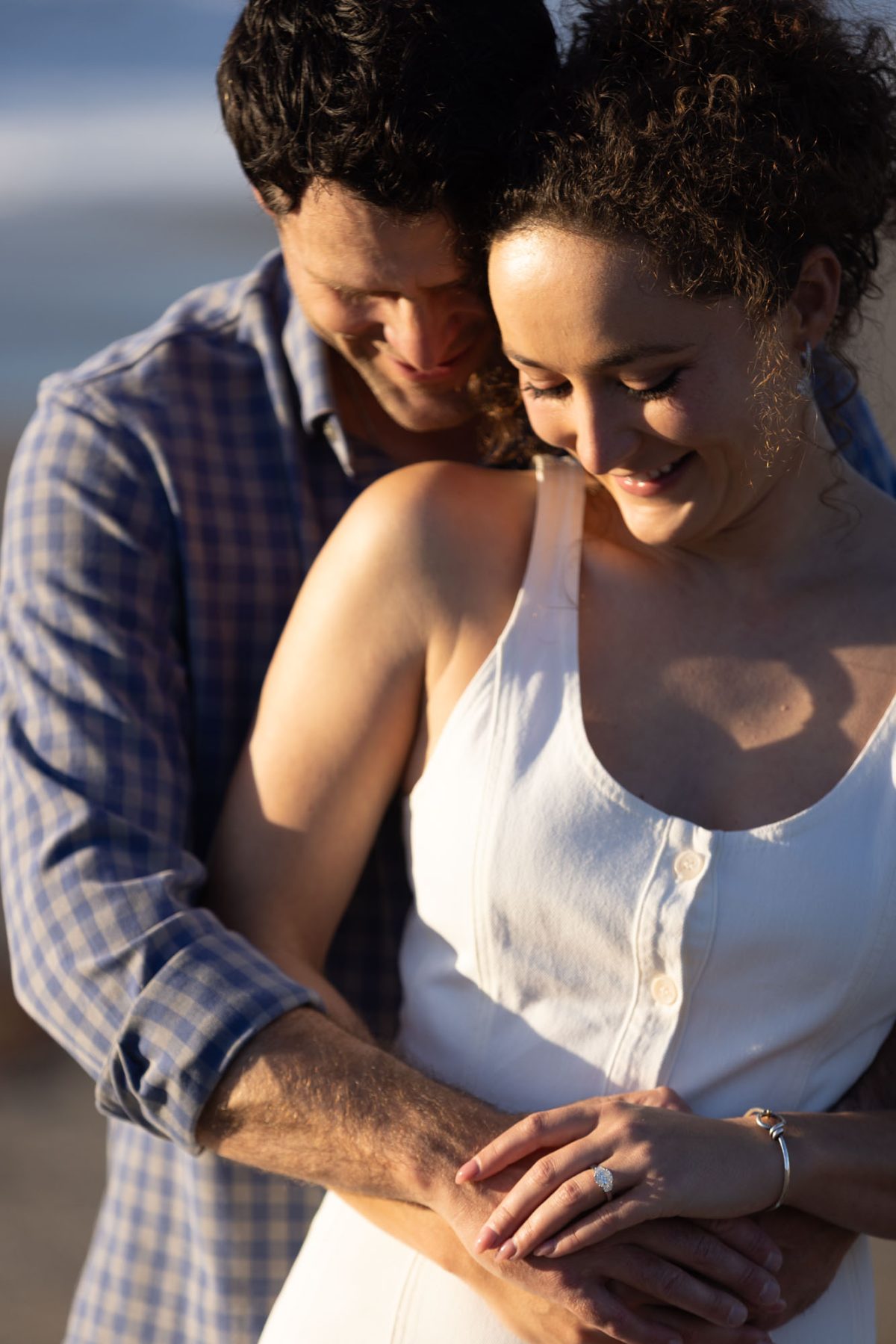 Intimate close-up of a joyful couple during their engagement photos in Bodega Bay, the man with dark curly hair in a blue plaid shirt embracing the woman with curly updo in a white sleeveless dress, their faces close and smiling tenderly at each other, hands intertwined showcasing a sparkling oval diamond engagement ring on her finger, with a soft hazy blue ocean backdrop evoking romance and bliss.