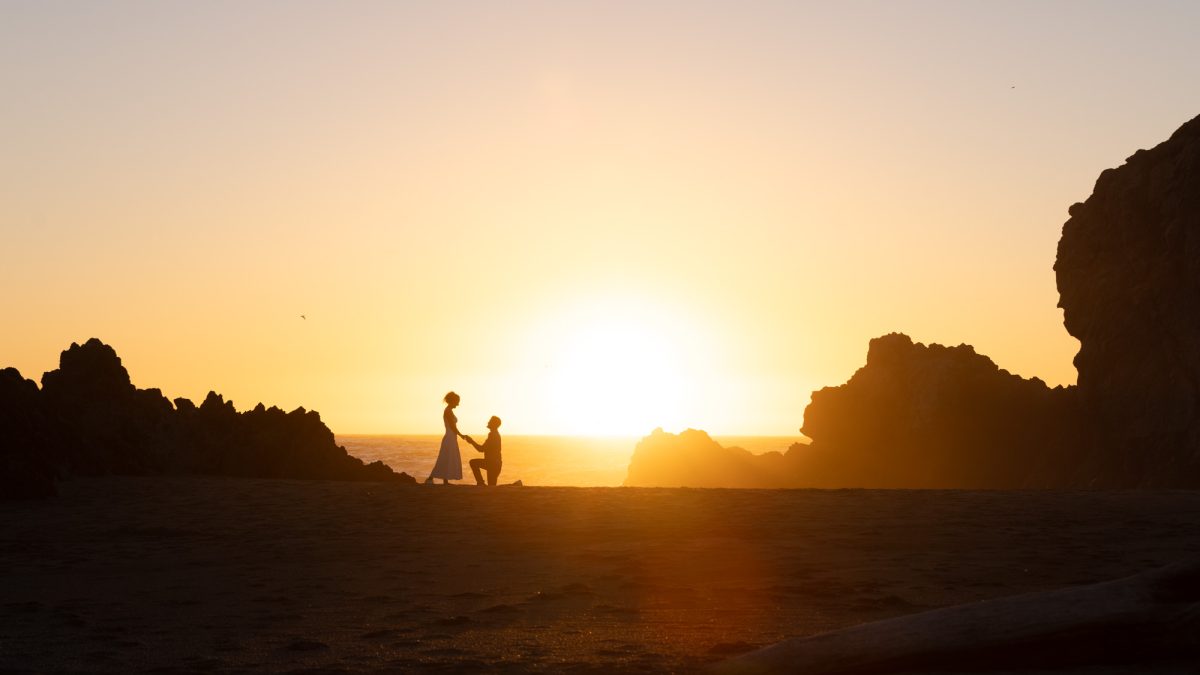 Bodega Bay surprise proposal captured in a striking silhouette against a radiant sunset over the Pacific Ocean. A man kneels on one knee on the dark sandy beach, tenderly holding the hand of a woman in a light flowing dress as she stands before him. Intense golden backlighting from the low sun creates a warm orange and yellow glow across the sky, with subtle lens flare and jagged rock formations framing the centered couple in this serene coastal composition.