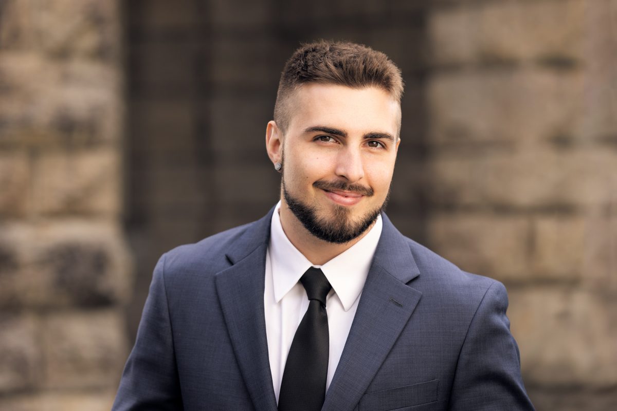 A young man stands confidently in front of a neutral stone building, wearing a tailored dark blue suit paired with a crisp white shirt and a black tie. His short, neatly trimmed hair and full beard are well-groomed, and he sports small stud earrings in both ears. With a subtle smile and direct eye contact, his expression conveys professionalism and approachability. The background is softly blurred, placing full focus on the subject while maintaining a sense of architectural context. This portrait is a polished example of Real Estate Headshots Sonoma County.