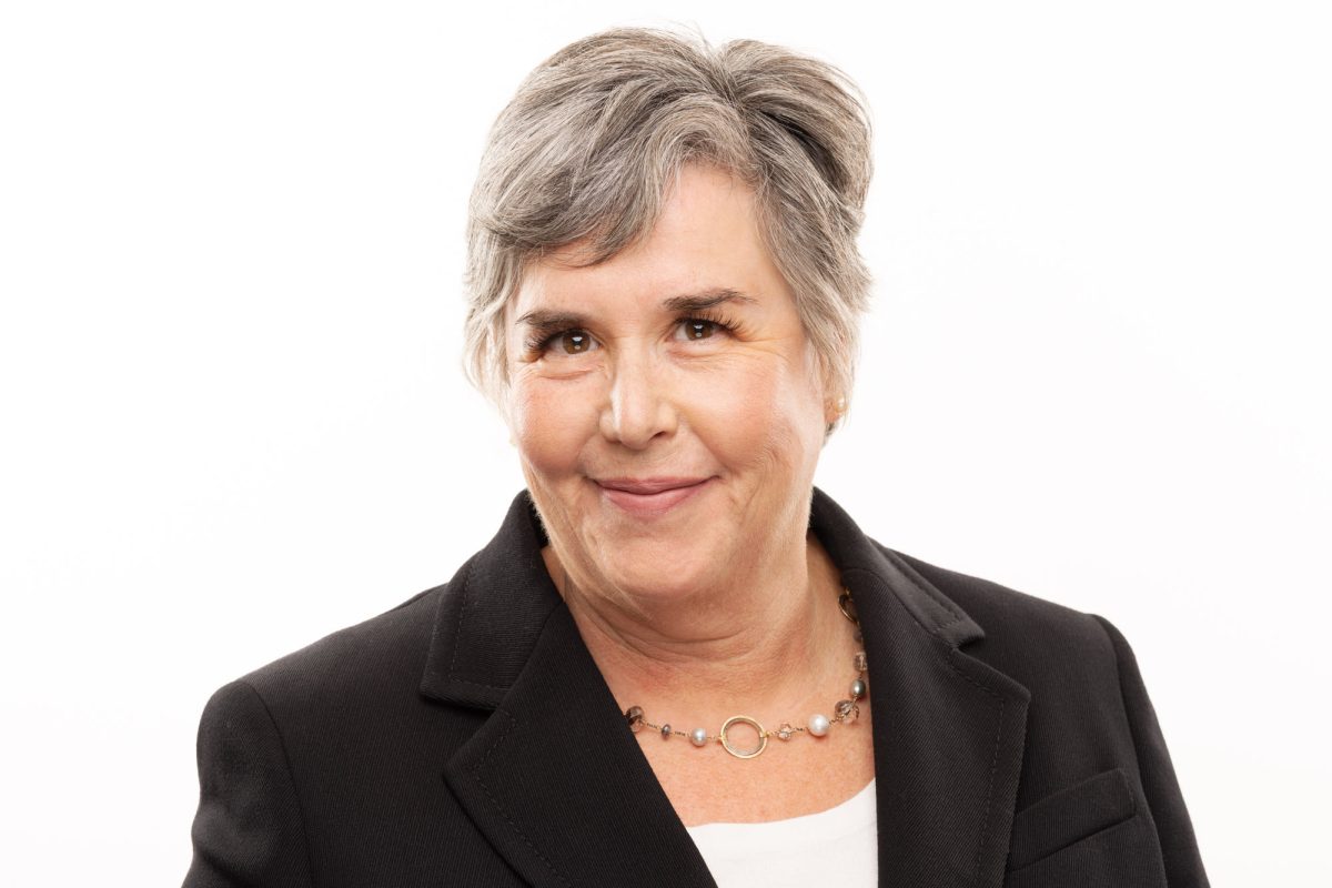 A professional woman stands against a clean white background, exuding warmth and confidence with a gentle smile. Her short, neatly styled gray hair frames her face, and her eyes are expressive and friendly. She wears a tailored black blazer over a white top, creating a classic and polished look. A delicate necklace featuring a mix of pearls and small gemstones with a gold ring centerpiece adds a subtle touch of elegance. The soft, even lighting highlights her features with clarity and professionalism, making this portrait a strong representation of Attorney Headshots Santa Rosa.