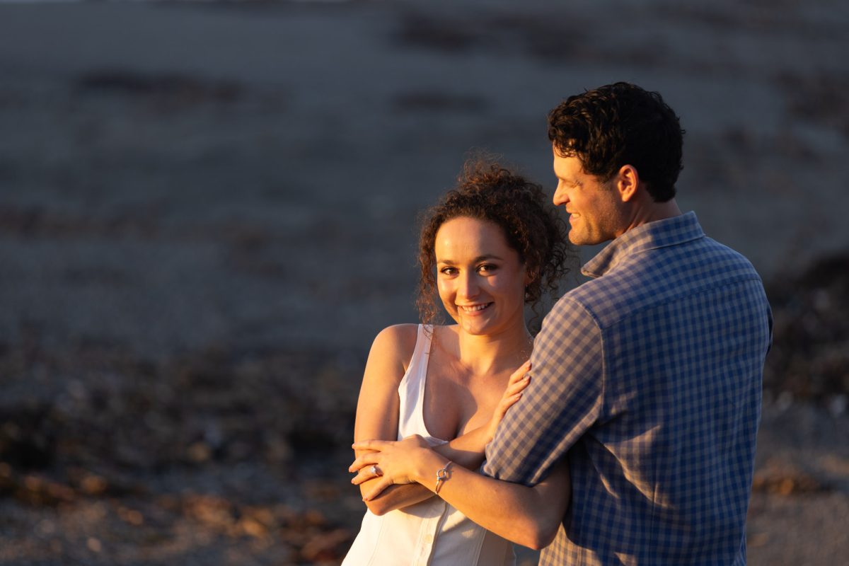 A woman with curly brown hair in a white top smiles at the camera while standing close with her partner during authentic couples portraits in Bodega Bay. The man with dark curly hair wearing a blue plaid shirt wraps his arm around her as warm golden sunlight illuminates their joyful moment on the sandy beach.