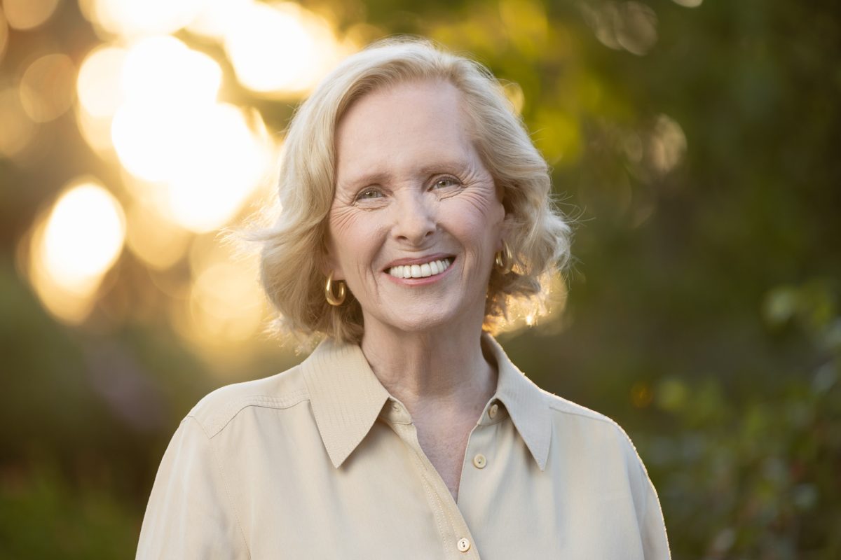 A woman with short, softly curled blonde hair stands outdoors during golden hour, bathed in the warm glow of sunlight filtering through blurred foliage in the background. She wears a light beige button-up shirt with a structured collar and gold hoop earrings that catch the soft light. Her broad smile and relaxed expression exude warmth and approachability. The backlighting gently outlines her hair, adding a luminous touch to the portrait. This radiant and natural image shows the impact author headshots can have on marketing.
