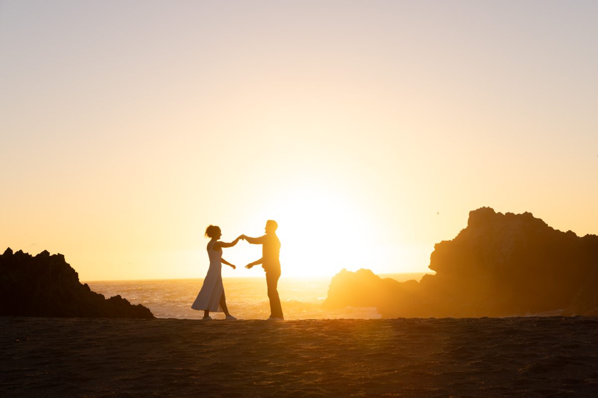 A woman with curly brown hair in a white dress holds hands with her partner during this beautiful Sonoma county surprise proposal. The man with dark curly hair stands facing her as warm golden sunlight creates a dramatic silhouette of their moment on the sandy beach with the ocean and large rock formations behind them.