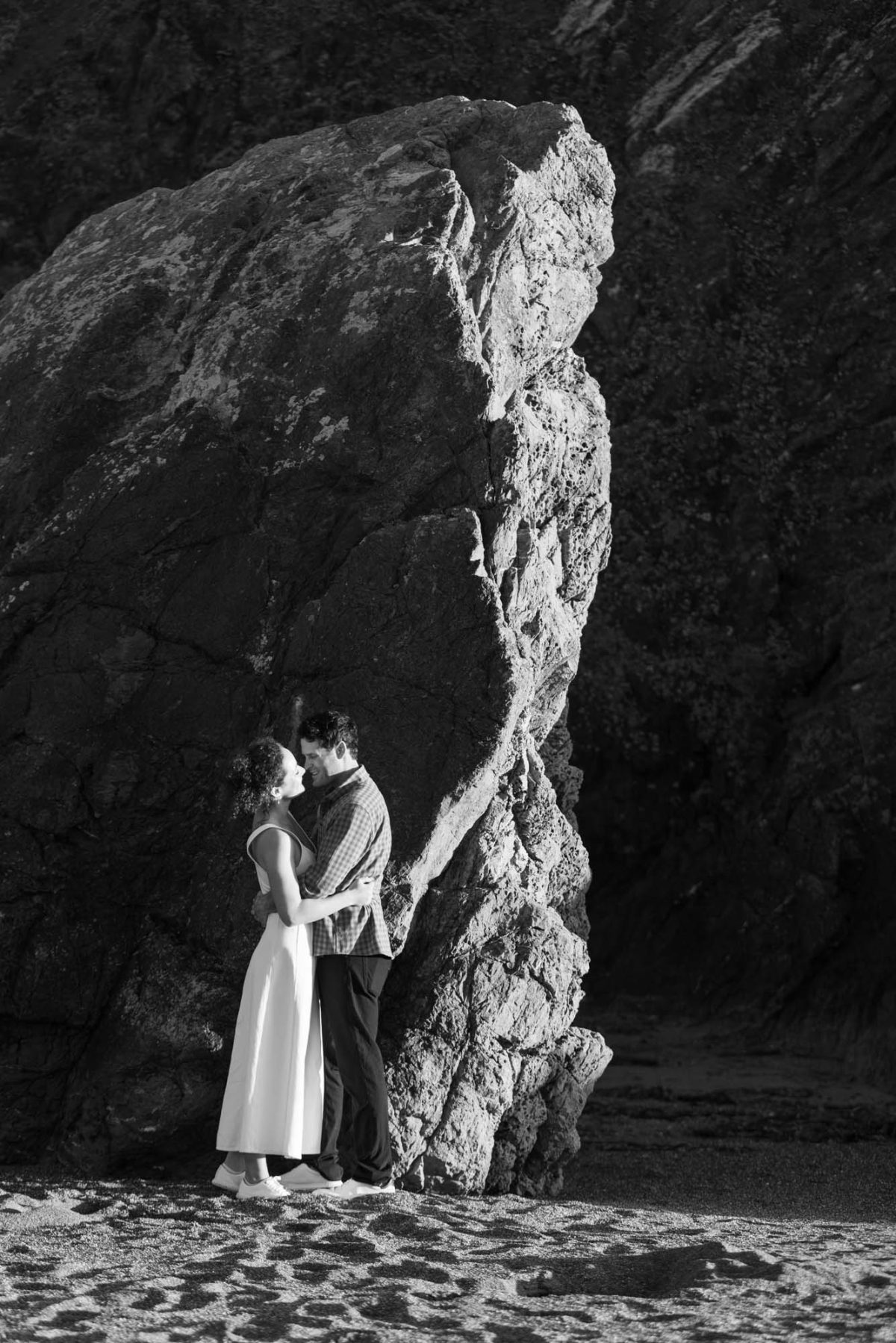 A woman with curly hair in a white sleeveless dress embraces a man wearing a checkered shirt as they stand close together gazing at each other beside a massive rock formation on the sandy beach. The black and white photograph captures dramatic natural lighting and strong shadows across the rocky landscape in their black and white beach engagement photos along the bodega bay coastline.