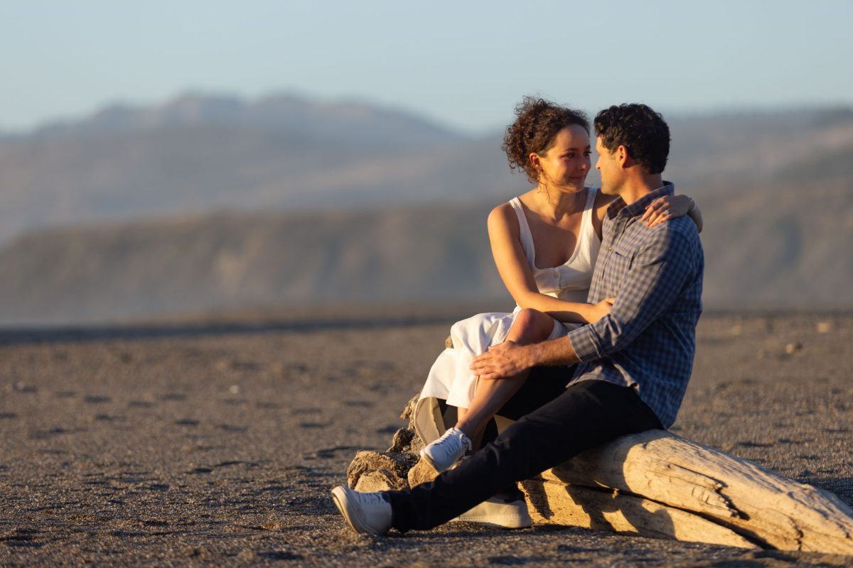 A woman with curly brown hair in a white top sits closely with her fiance on a large driftwood log during their Bodega Bay, California engagement photo shoot. The man with dark curly hair wearing a blue plaid shirt has his arm around her as they look at each other while warm golden sunlight illuminates their close moment on the sandy beach.