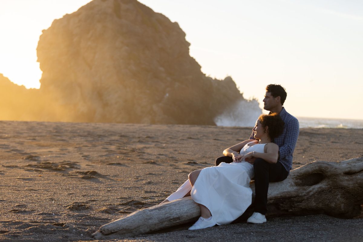 A woman with curly brown hair in a white dress sits on a large driftwood log with her partner during their bodega bay couples photos at sunset. The man with dark curly hair wearing a blue plaid shirt sits behind her with his arm around her as they both look out towards the ocean and dramatic rock formation while warm golden sunlight illuminates their close moment on the sandy beach.