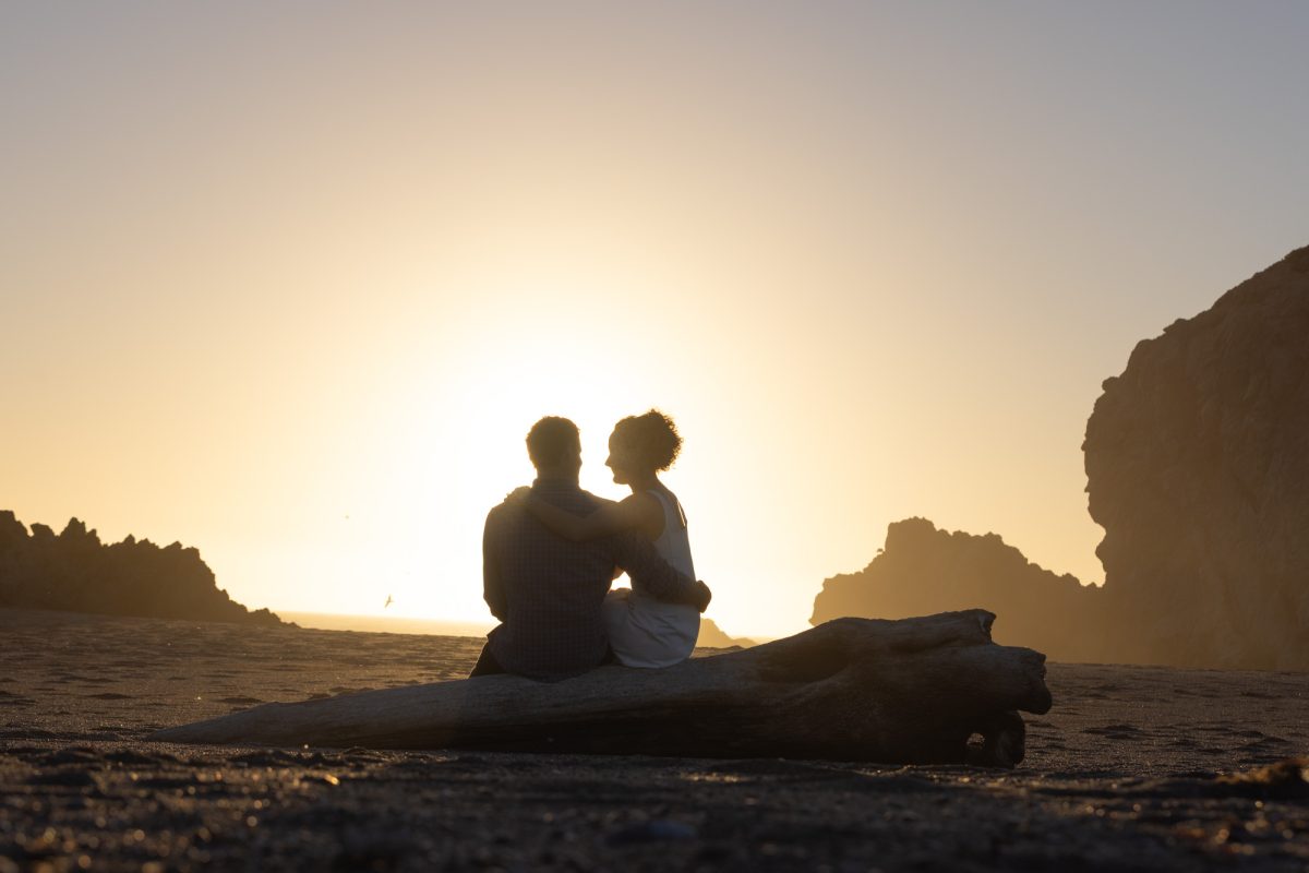 A woman with curly brown hair in a white top sits closely with her partner on a large driftwood log during a bodega bay engagement session. The man with dark curly hair wearing a plaid shirt has his arm around her as they look at each other while warm golden sunlight creates a dramatic silhouette of their close moment on the sandy beach with large rock formations behind them.