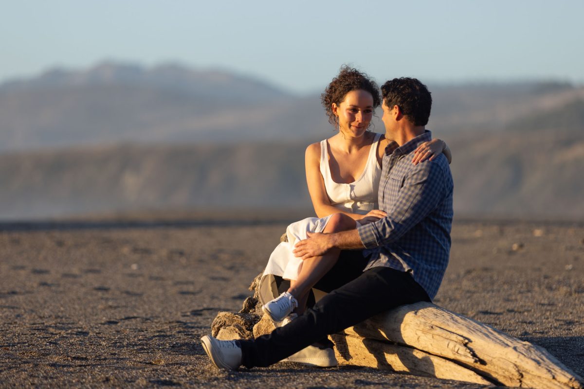 A woman with curly brown hair in a white top sits closely with her partner on a large driftwood log during a Bodega Bay engagement photo shoot. The man with dark curly hair wearing a blue plaid shirt has his arm around her as they look at each other with smiles while warm golden sunlight illuminates their close moment on the sandy beach.