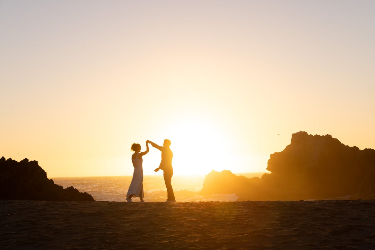 A woman with curly brown hair in a white dress holds her partner's hand high as they dance together after their Bodega bay surprise proposal pictures. The man with dark curly hair faces her while warm golden sunlight creates a dramatic silhouette of their moment on the sandy beach with the ocean and large rock formations behind them.